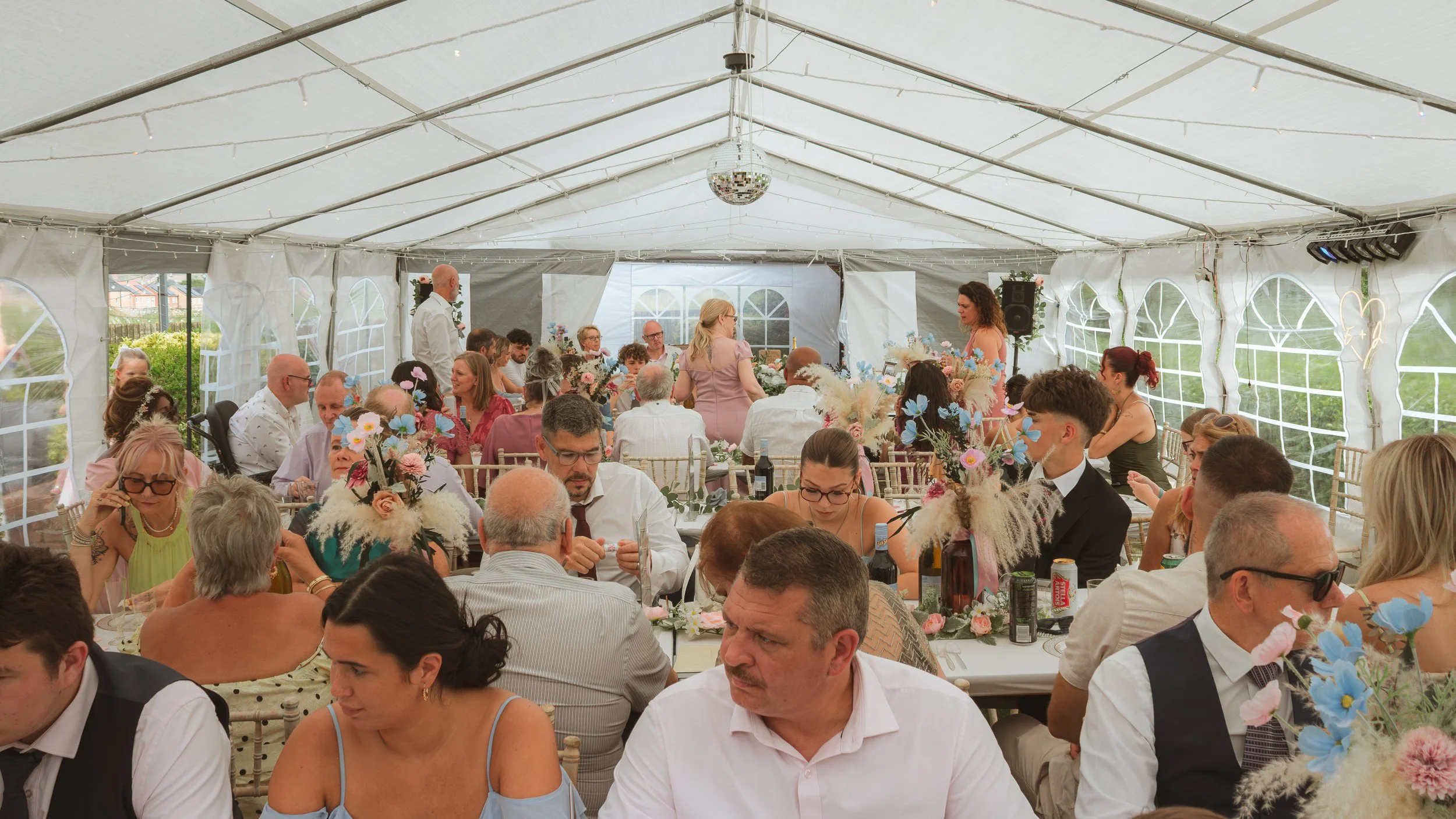 A large group of people sitting at long decorated tables inside a white event tent during a wedding reception or celebration.
