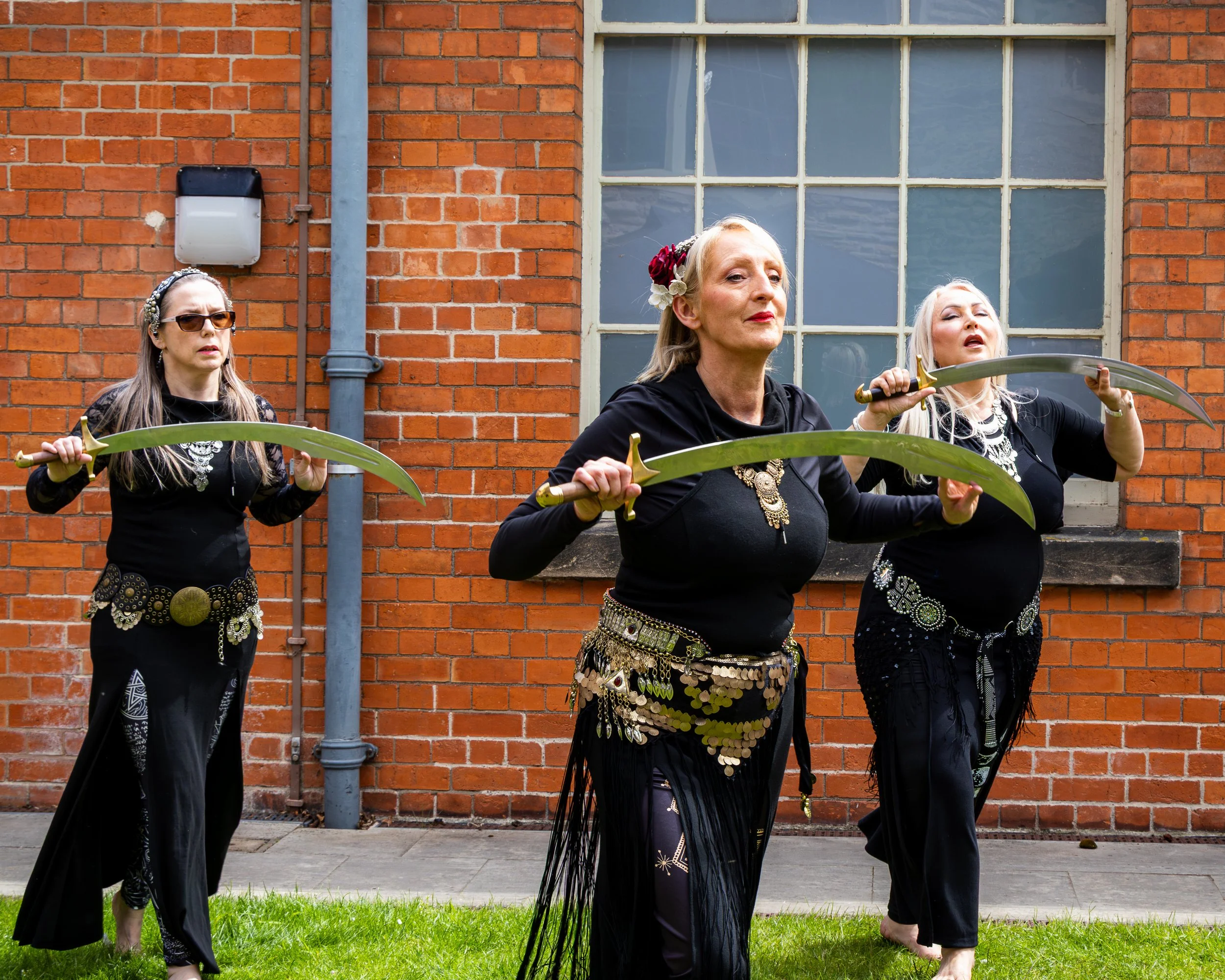 Three women dressed in black traditional attire standing outdoors in front of a red brick building, holding curved swords.