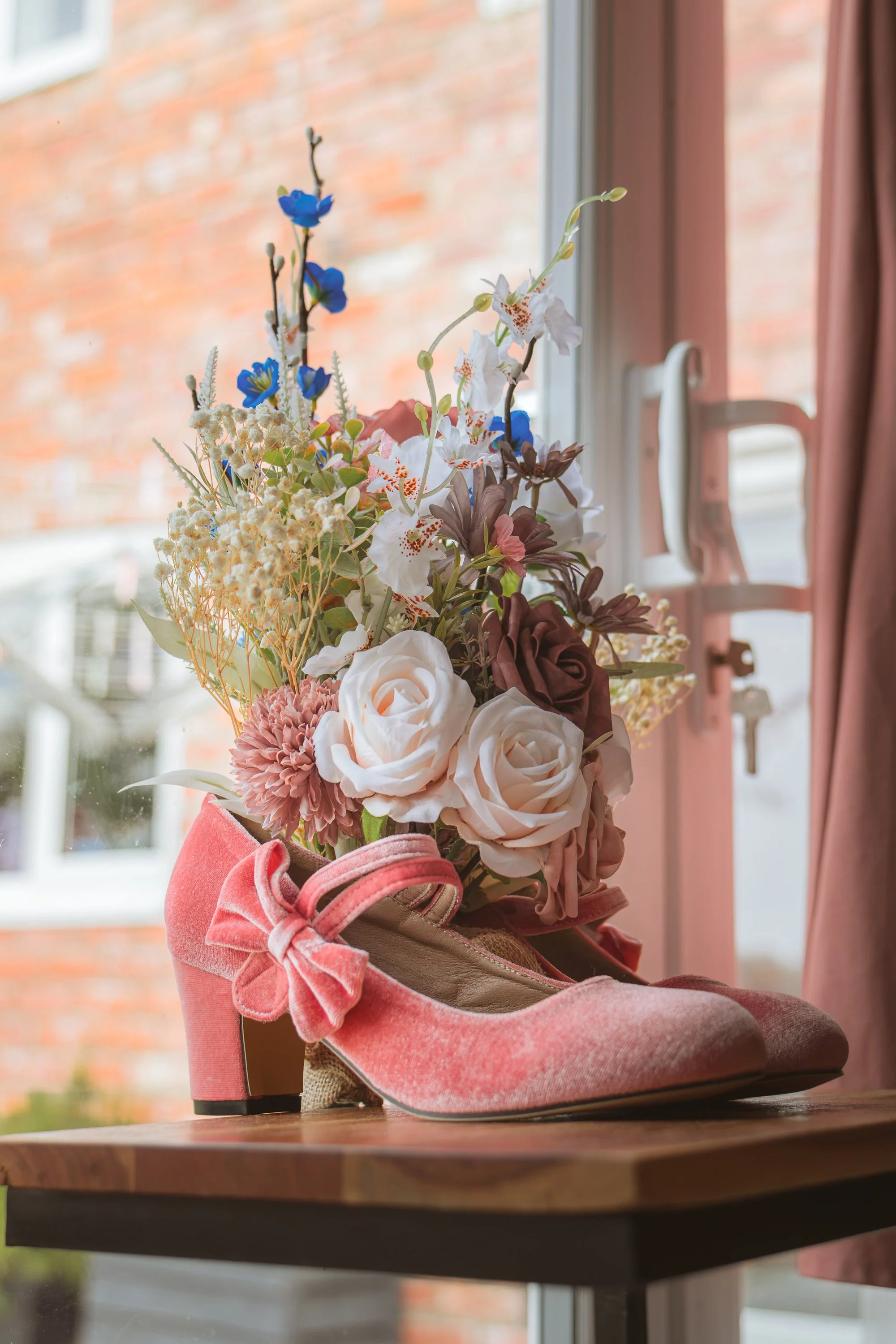 Pink high-heeled shoe with a curly bow on the toe, filled with a bouquet of white, pink, and blue flowers, placed on a wooden table near a window with a brick wall visible outside.