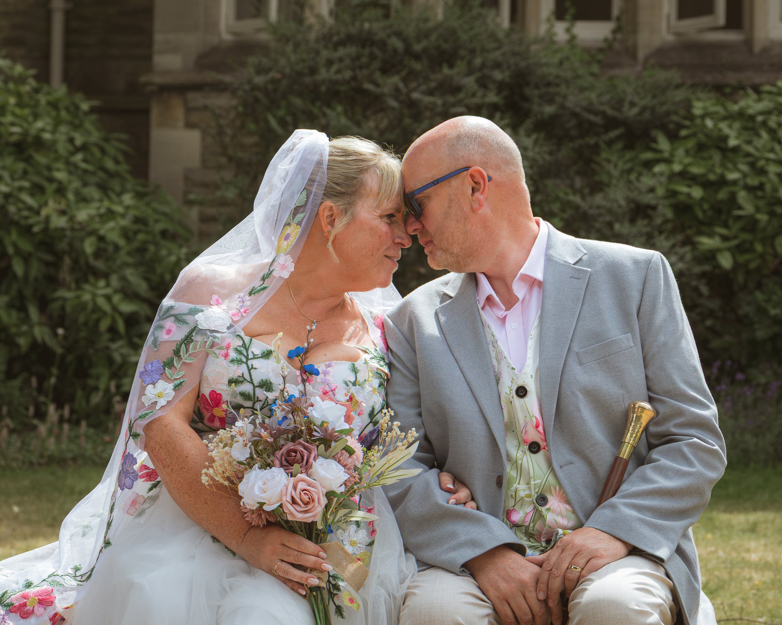 A bride and groom sitting close with their foreheads touching outdoors, surrounded by greenery, during a wedding ceremony. The bride is holding a bouquet of flowers, wearing a dress with floral embroidery and a veil. The groom is dressed in a light g