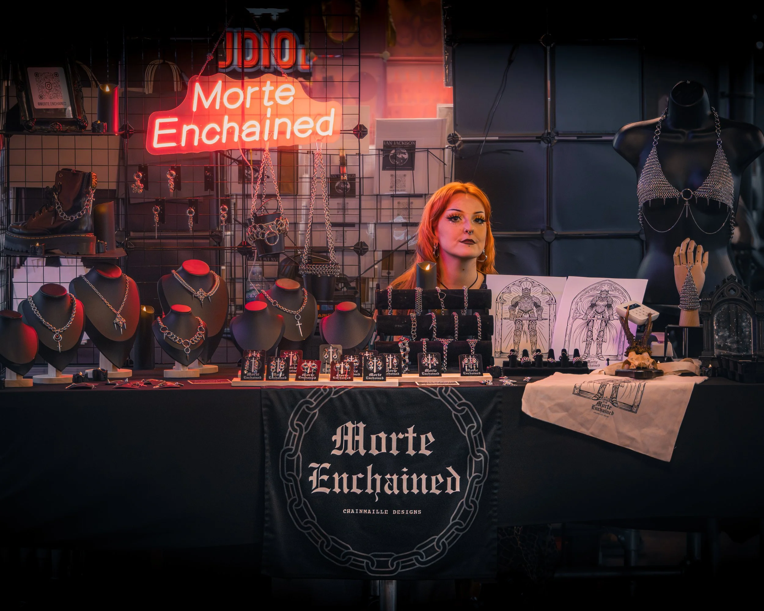 Jewelry and accessories displayed at a market stall with a neon sign that reads 'Morte Enchained.'