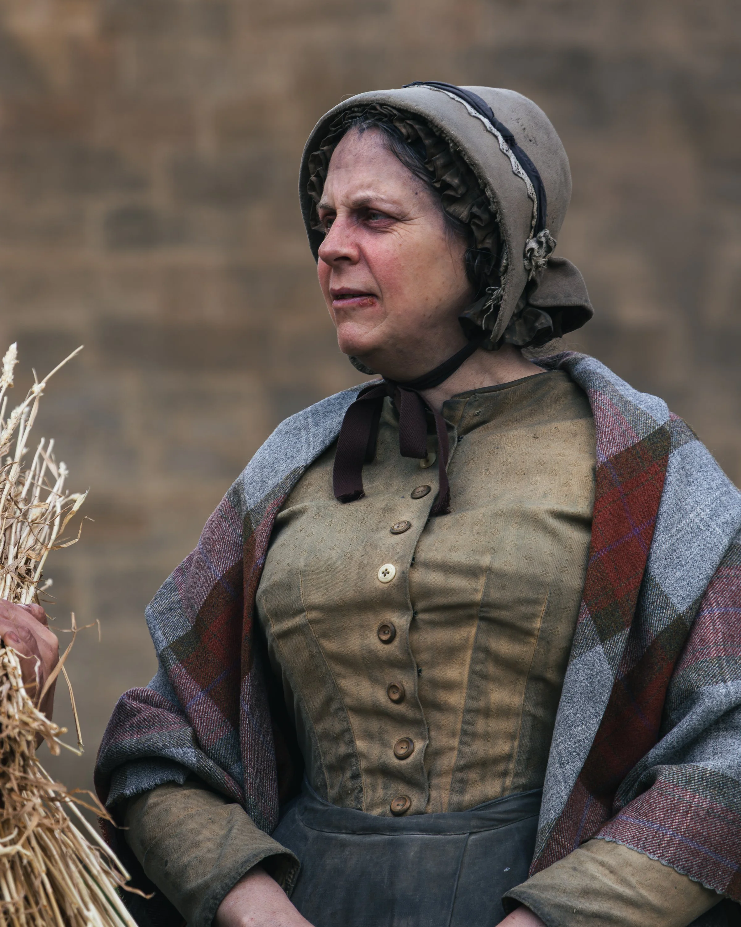 Close-up of a woman dressed in rustic, vintage clothing, including a bonnet, layered coat, and apron, standing outdoors with a blurred background of dry grass and earth.