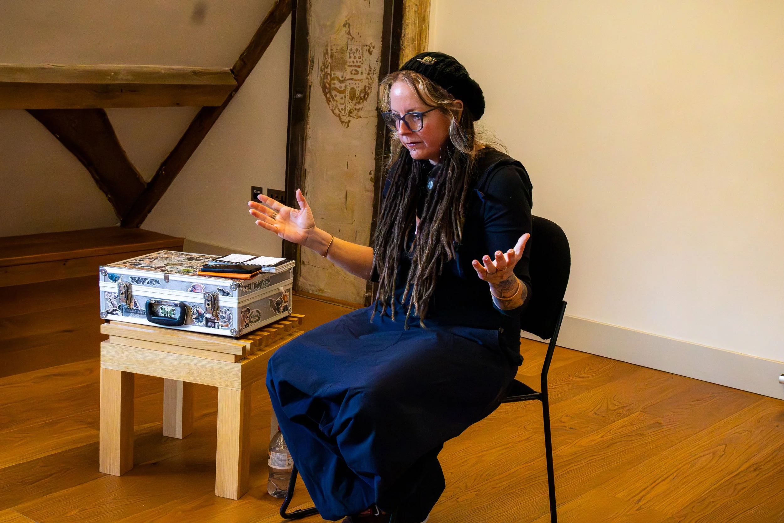 Woman with long dreadlocks, glasses, and a black beret sitting at a wooden table, speaking or gesturing with hands, with a beige wall and artwork behind her, and a colorful decorated suitcase on the table.