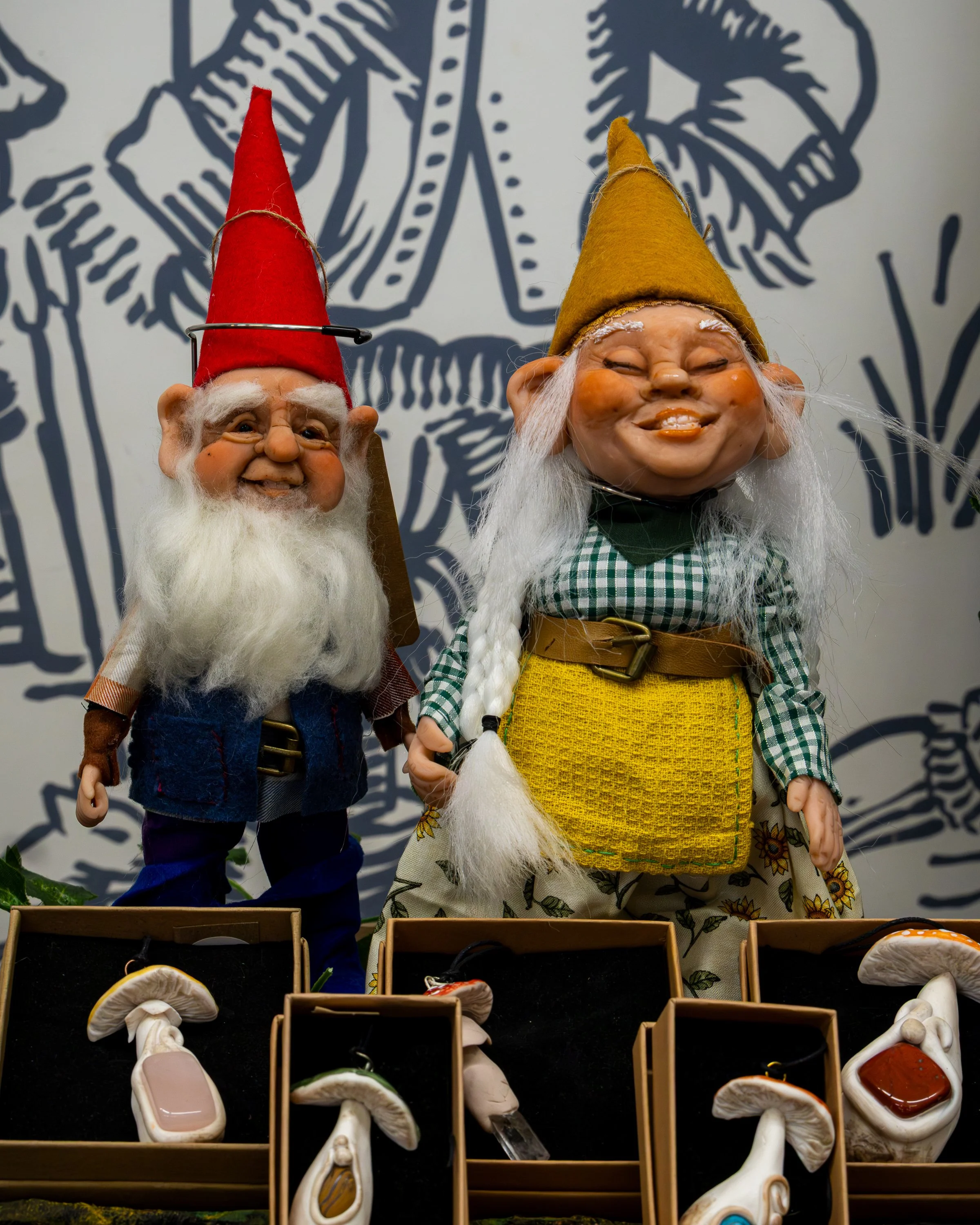 Two elf figurines with pointy hats, one red and one yellow, standing behind a display of various mushrooms in small wooden boxes. The background features a blue outline drawing of an elf figure.