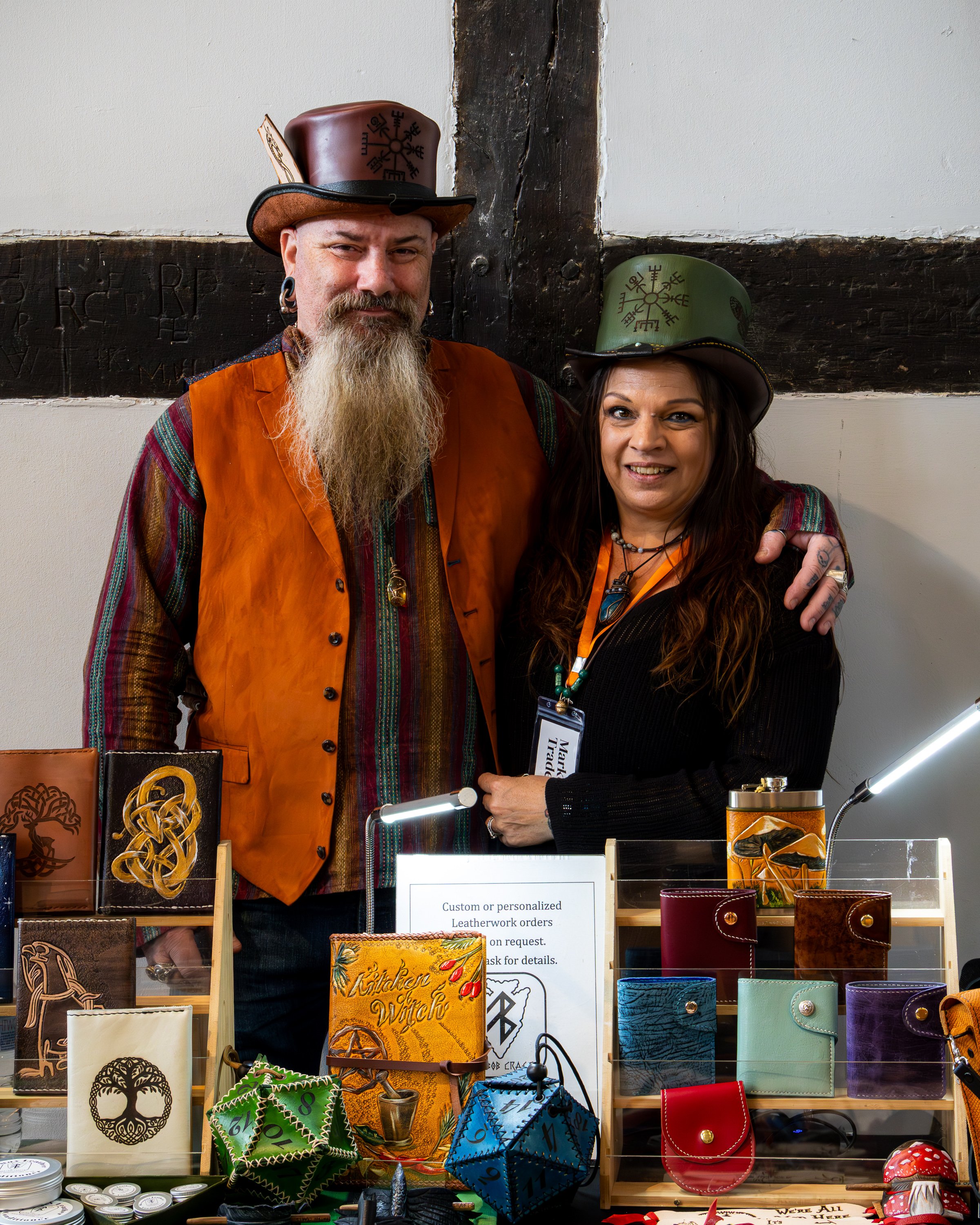 Two people dressed in costume at a leatherwork and craft stall. The man has a long beard, earrings, and is wearing a hat with rune symbols, an orange vest, and a plaid shirt. The woman has long dark hair, makeup, and is wearing a hat with rune symbol