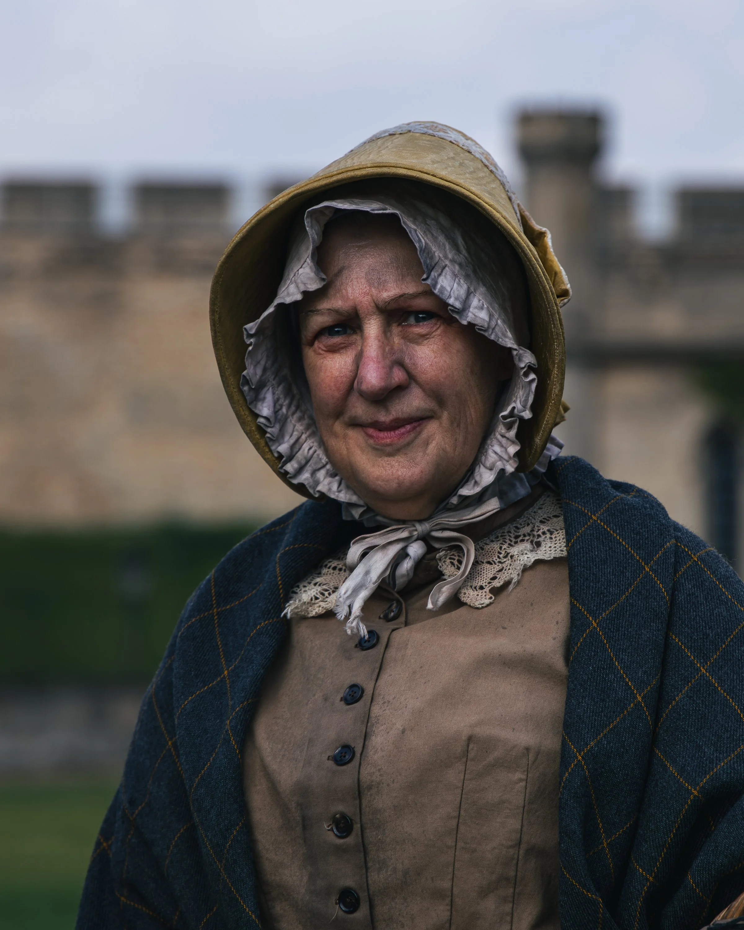A woman dressed as a beekeeper, wearing a hooded protective suit with a hat, scarf, and a dark blue jacket with yellow checkered pattern, standing outdoors with a blurred castle or large building in the background.