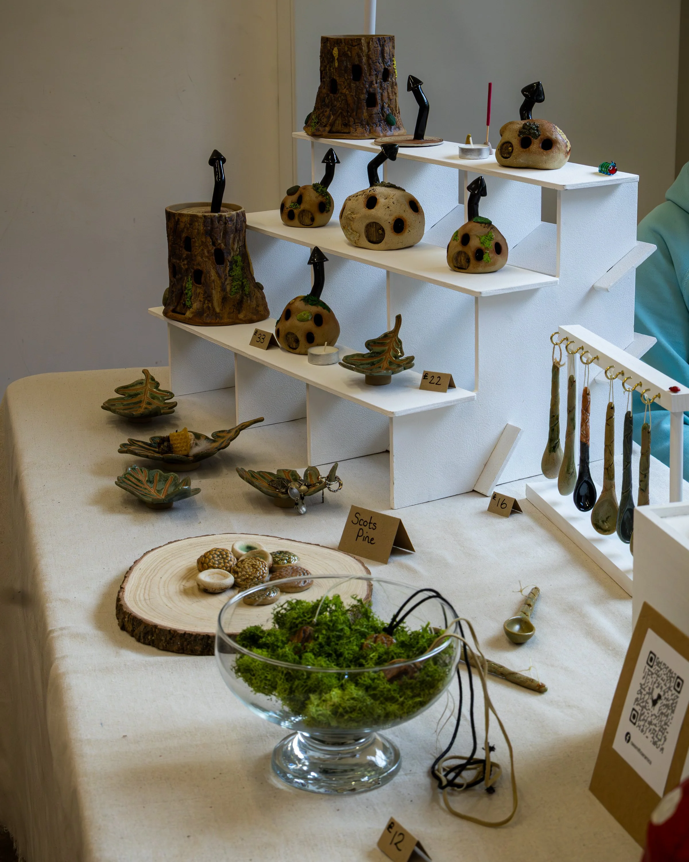Display of various handcrafted ceramic and wood objects on white shelves, including figures resembling tree stumps and rocks with black mushroom-shaped protrusions, along with small bowls and decorative items, labeled with prices.