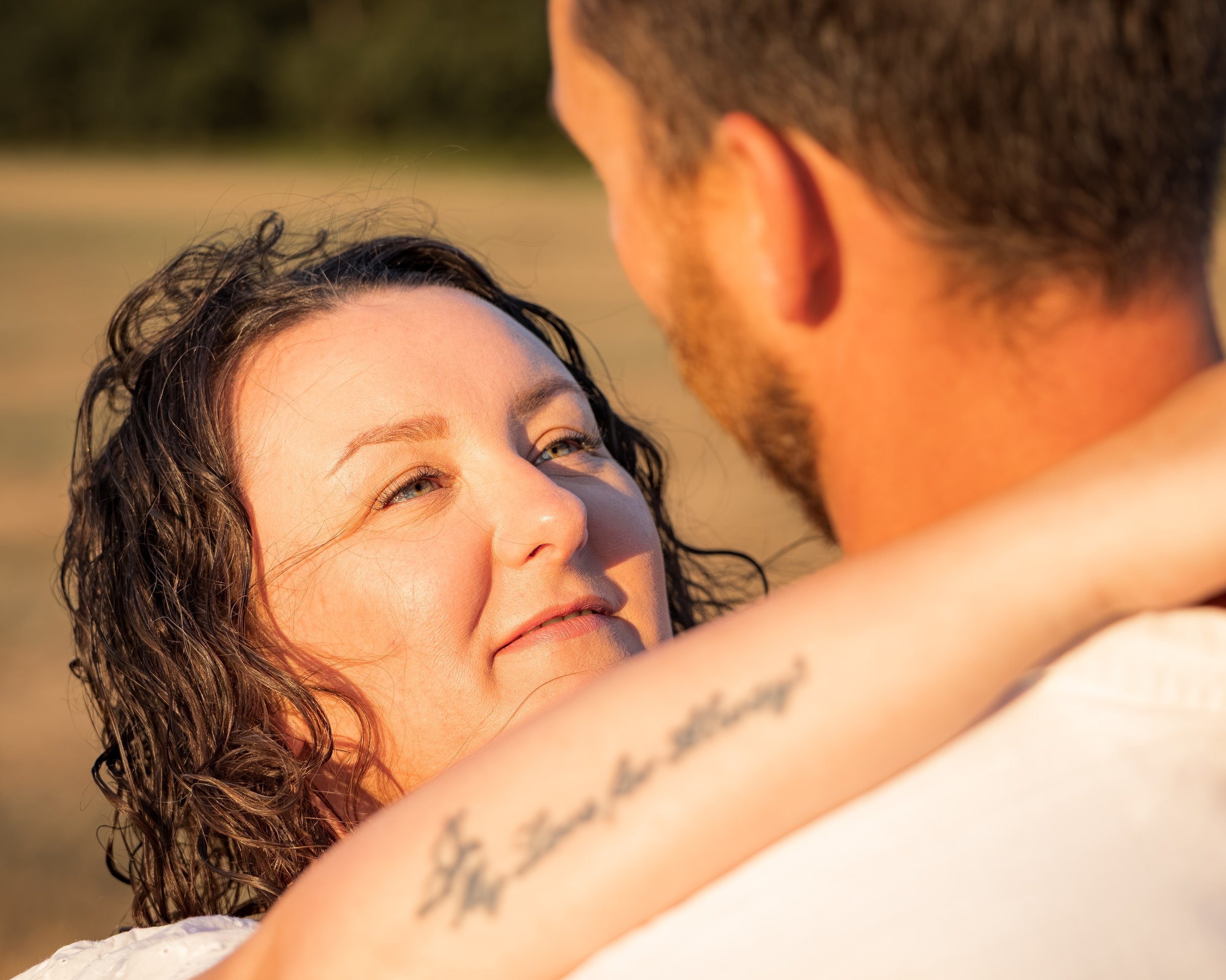 A woman with curly hair looks at a man with short hair and tattoos as they embrace outdoors during sunset.