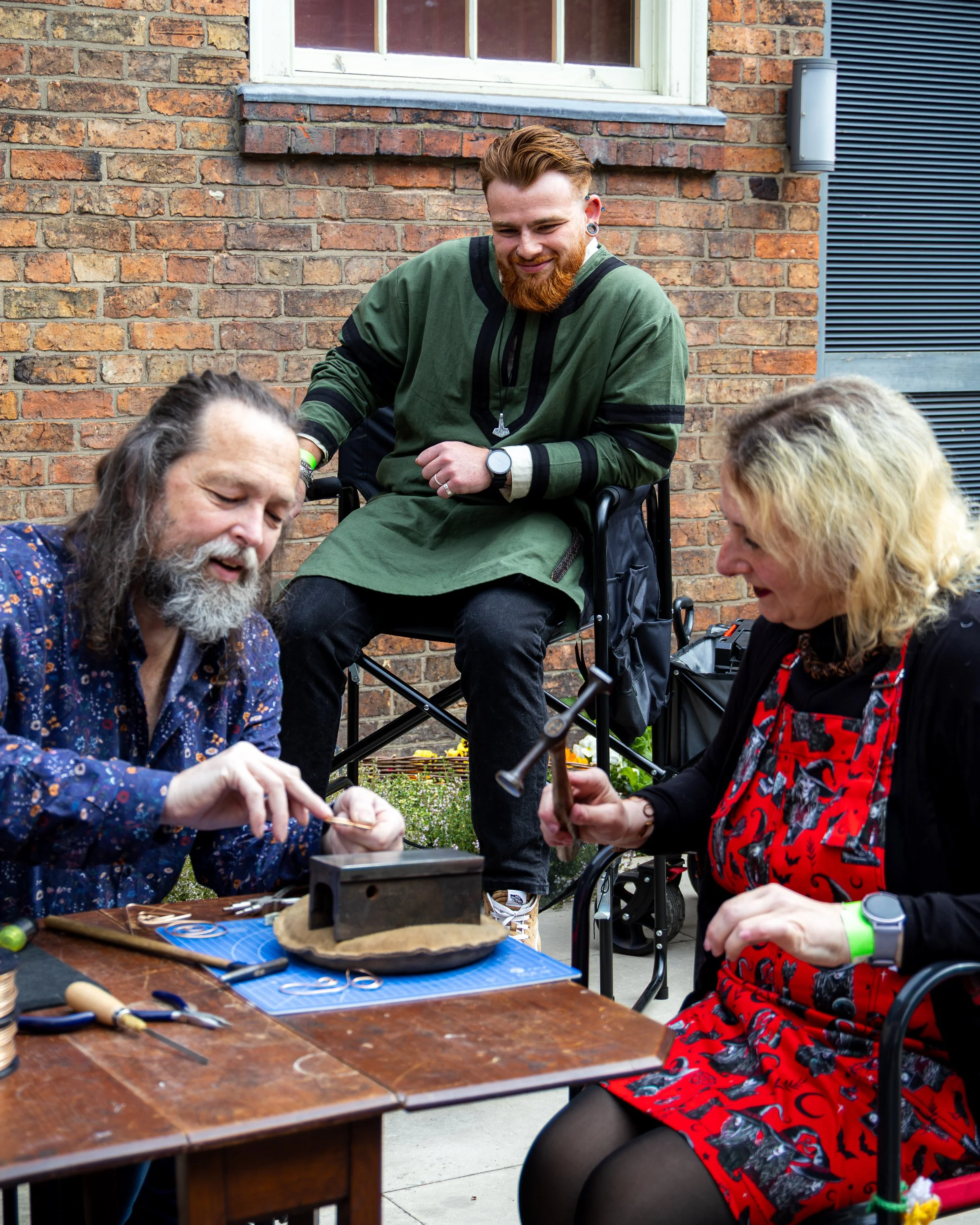 A group of people gathered outdoors next to a brick wall, working on a jewelry project on a wooden table, with tools and materials scattered on it. One person is seated in a wheelchair, smiling and observing others, who appear engaged in crafting or 