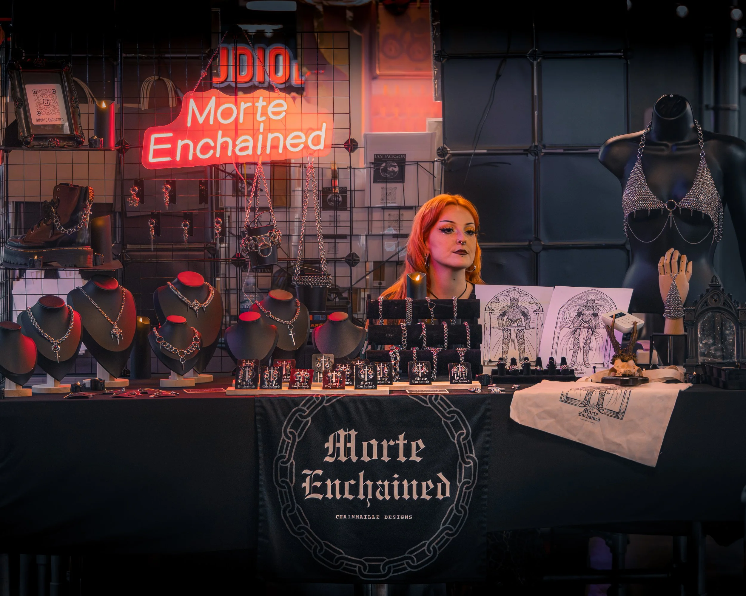 A display of Gothic and punk-style jewelry and accessories at a booth with a neon sign that reads 'Morte Enchained.' The jewelry includes necklaces, chains, and earrings, accompanied by sketches and artwork on the table. There is a mannequin torso ad