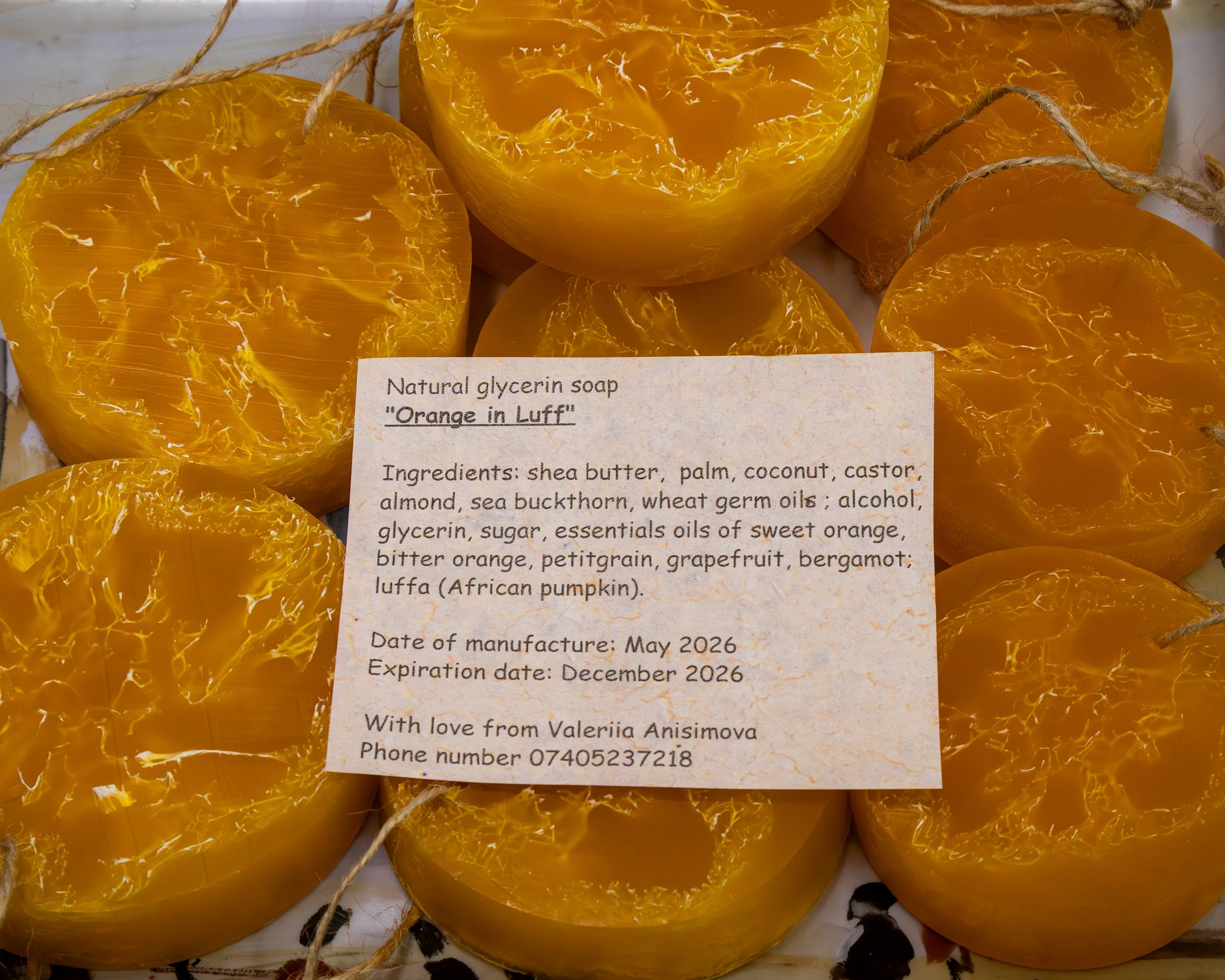 Natural glycerin soap labeled "Orange in Luff" with cut pieces showing orange-colored soap with intricate patterns, tied with twine, and a tag listing ingredients and manufacturing details.