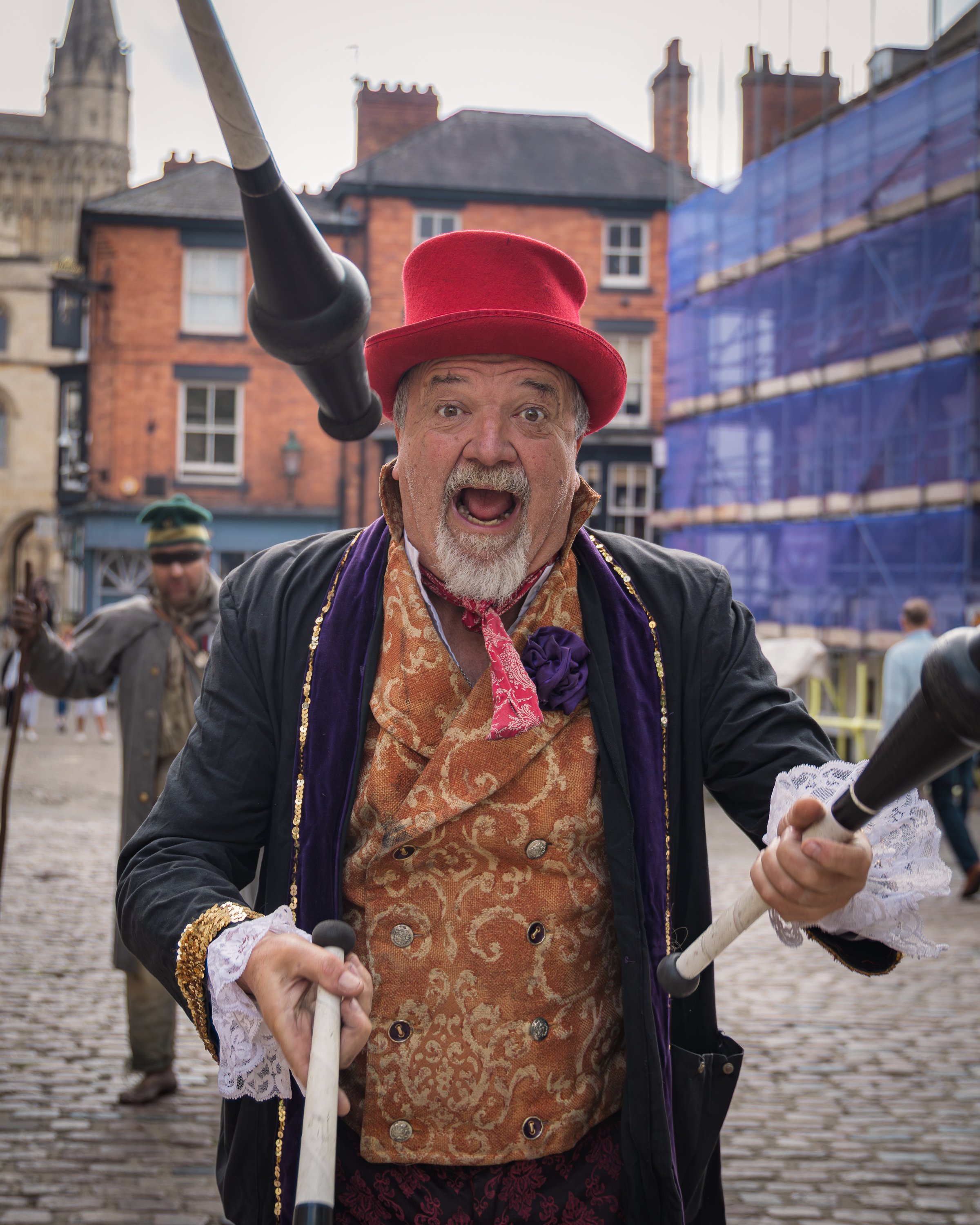 Man wearing a red top hat and ornate costume blowing a horn during a street festival.
