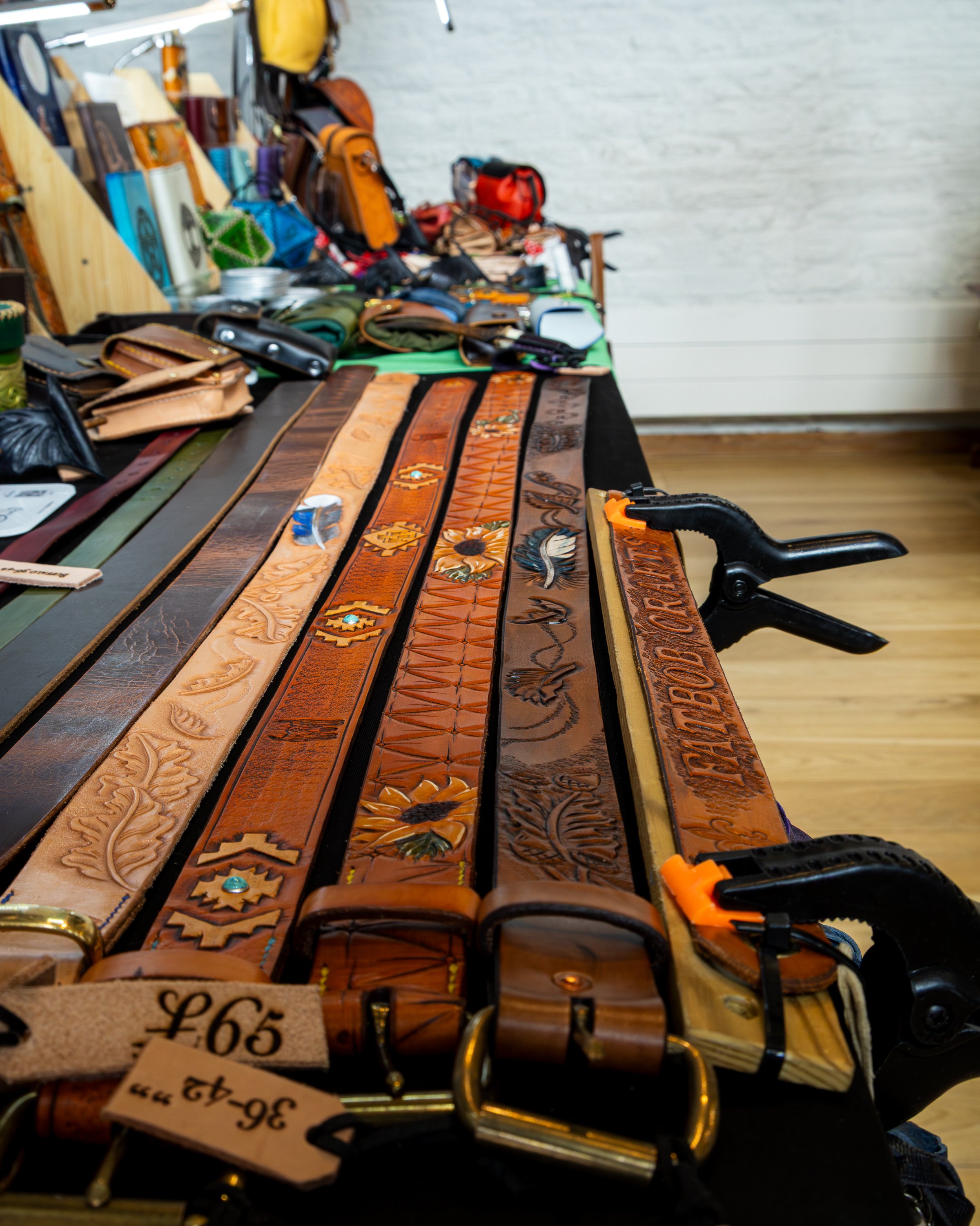 A table displaying various leather belts with different colors, patterns, and carvings, and some other accessories, at a market or store.