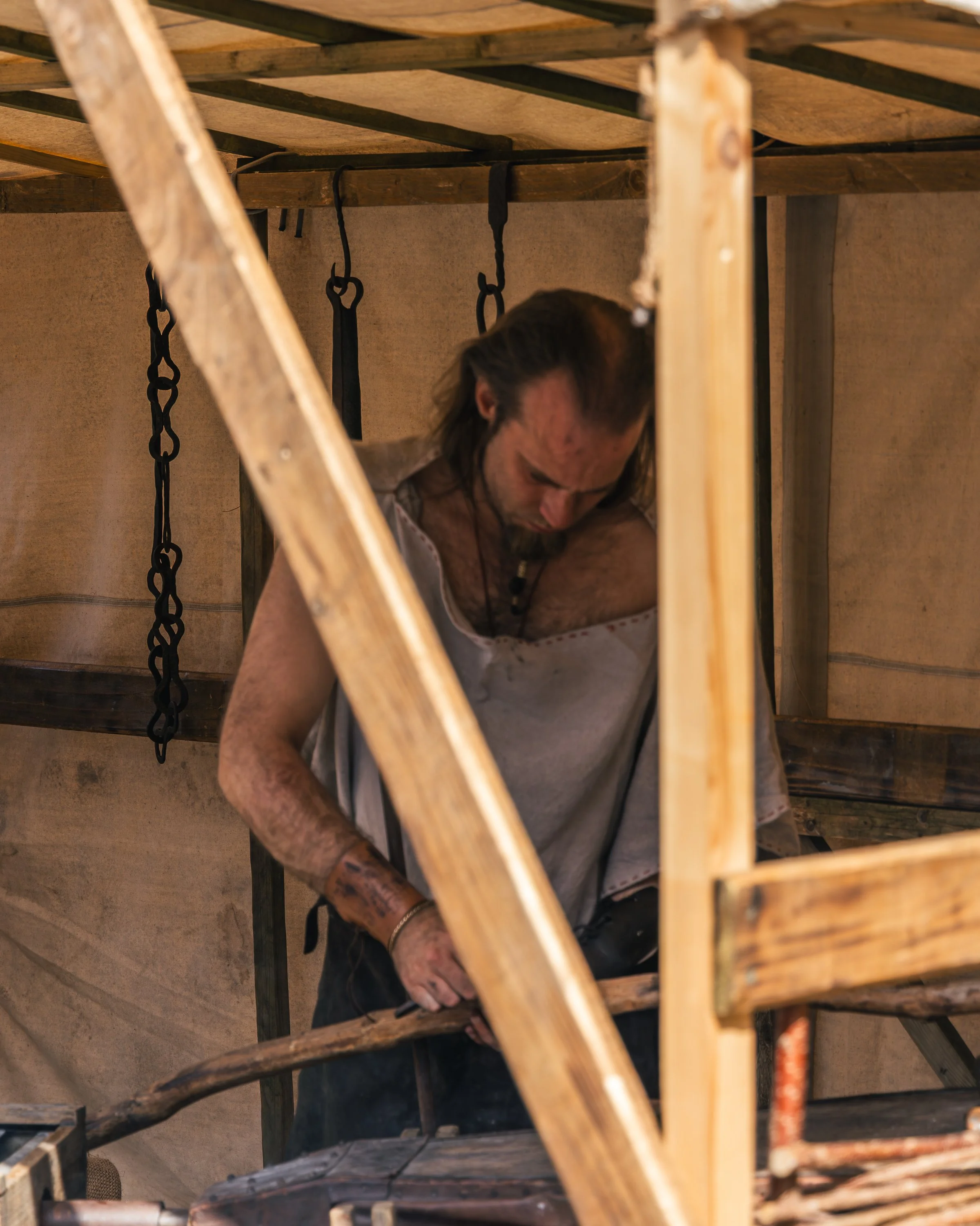 Man with long hair and tattoos sitting barefoot inside a wooden structure, with chains hanging from the ceiling.