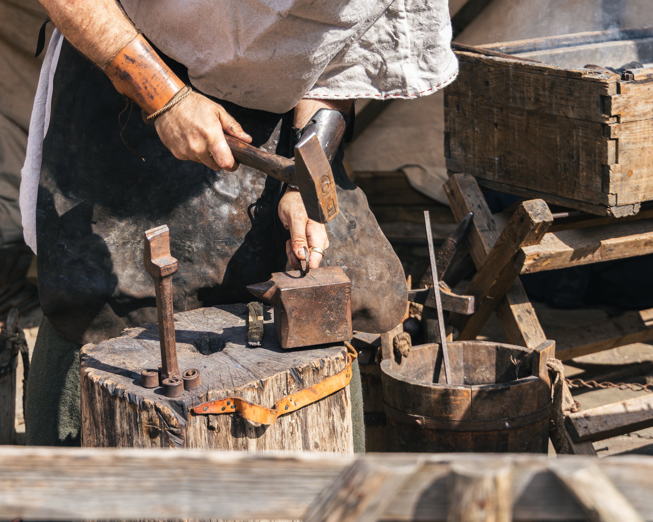 A person working on a woodworking project outdoors, wearing a light-colored shirt and dark apron, holding a hammer near a workbench with tools, wood, and wooden structures around.