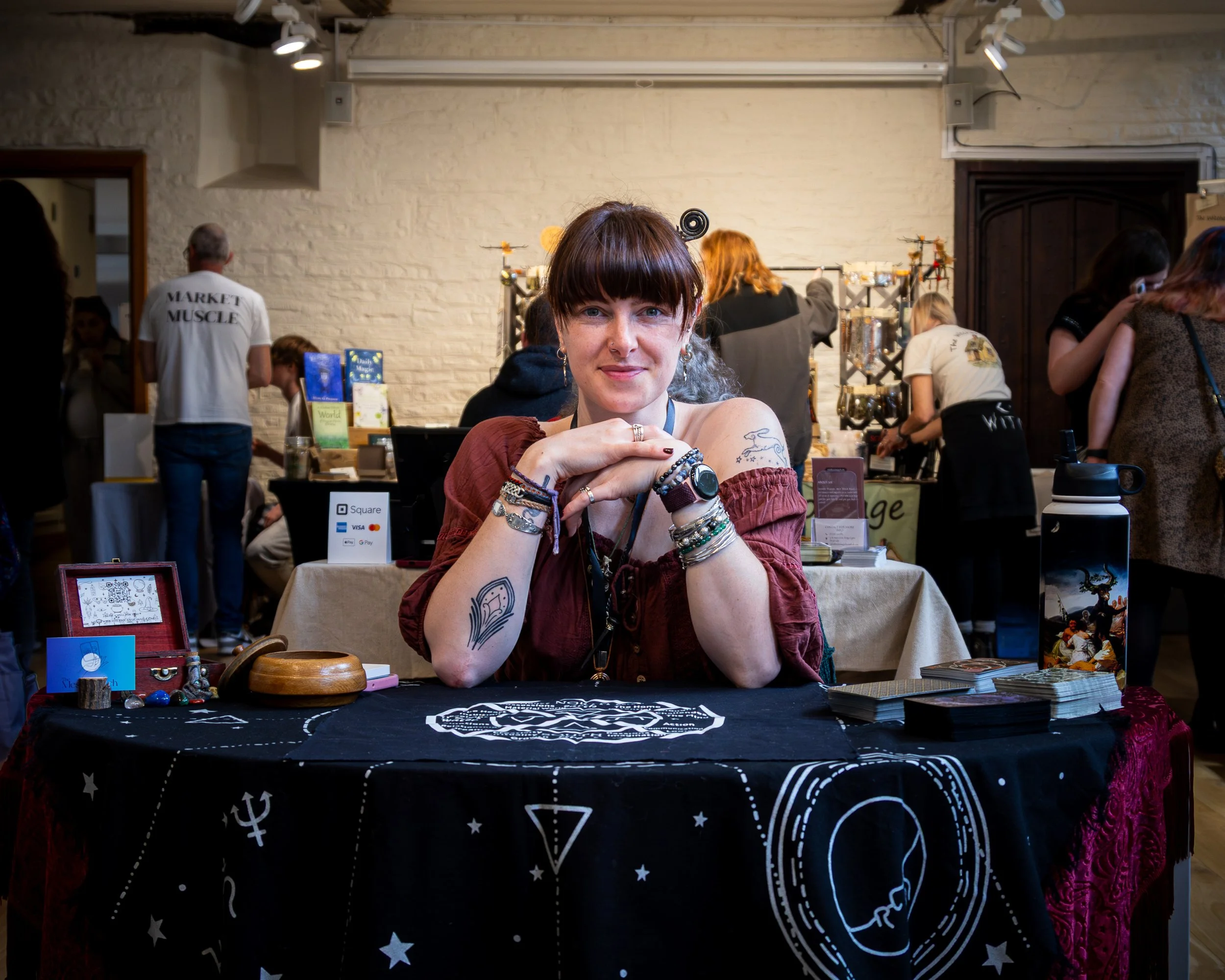A woman with tattoos and jewelry sitting at a booth with a black cloth featuring white space-themed symbols, smiling at the camera, with an indoor market or event in the background.