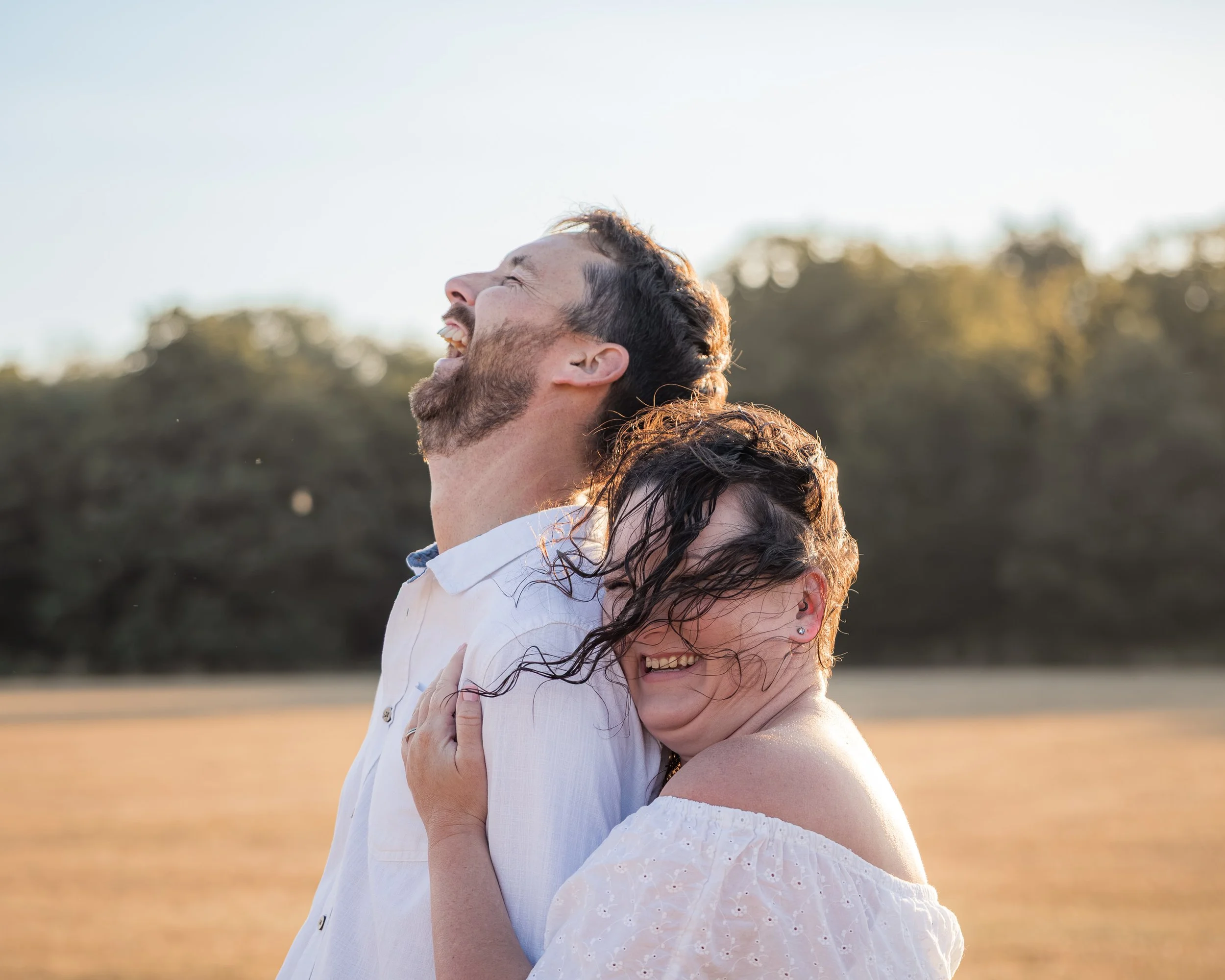 A happy couple embracing outdoors during sunset, smiling and laughing with their eyes closed.