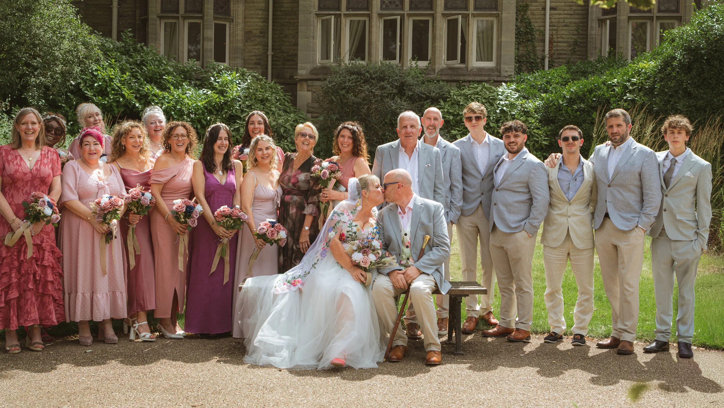 Group of people celebrating a wedding outdoors, with women holding bouquets, the bride and groom sitting in front, and a mix of men and women dressed in colorful formal attire.
