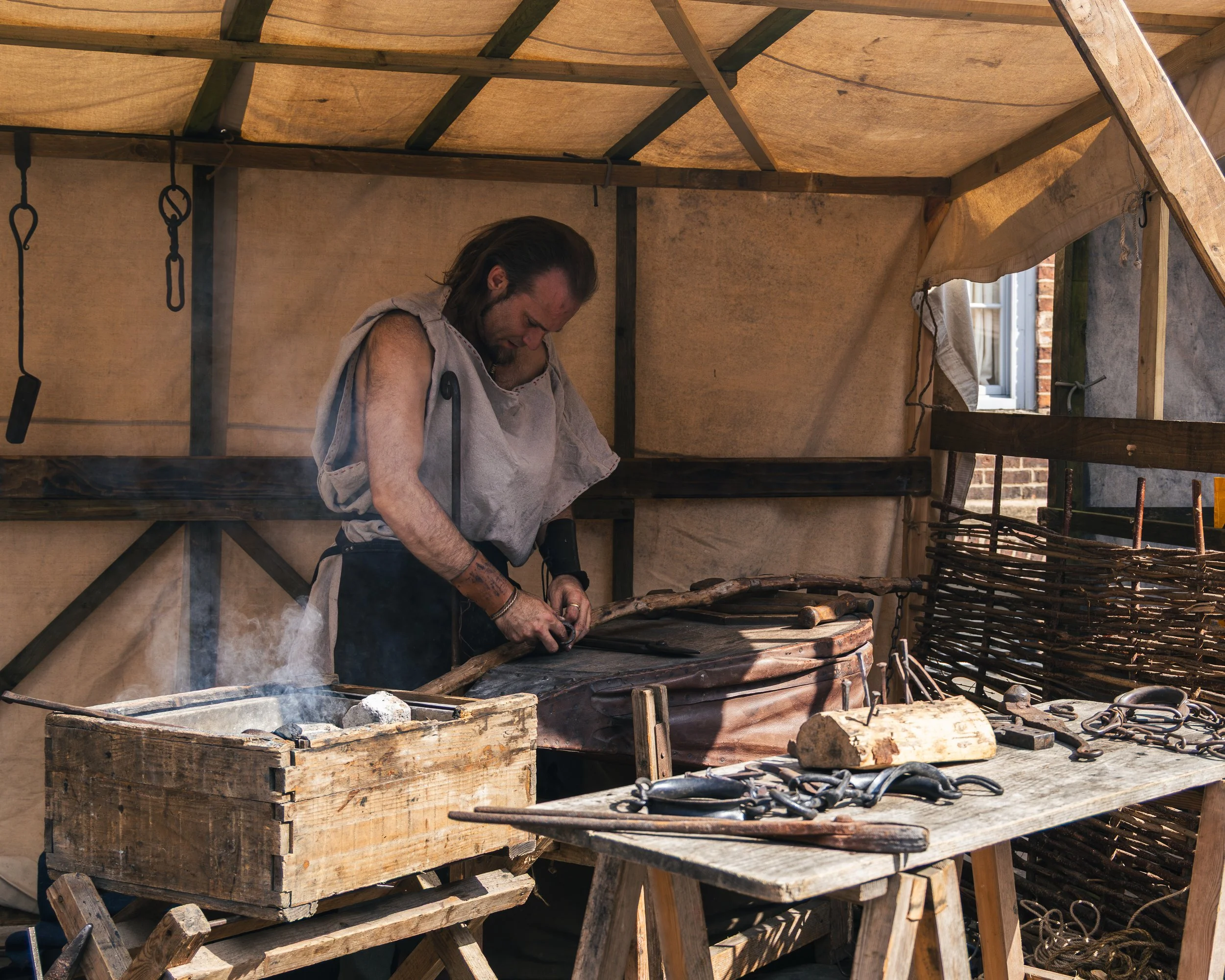 A man wearing medieval-style clothing working with wood inside a small open-air workshop with wooden and canvas structure and tools hanging from the ceiling.