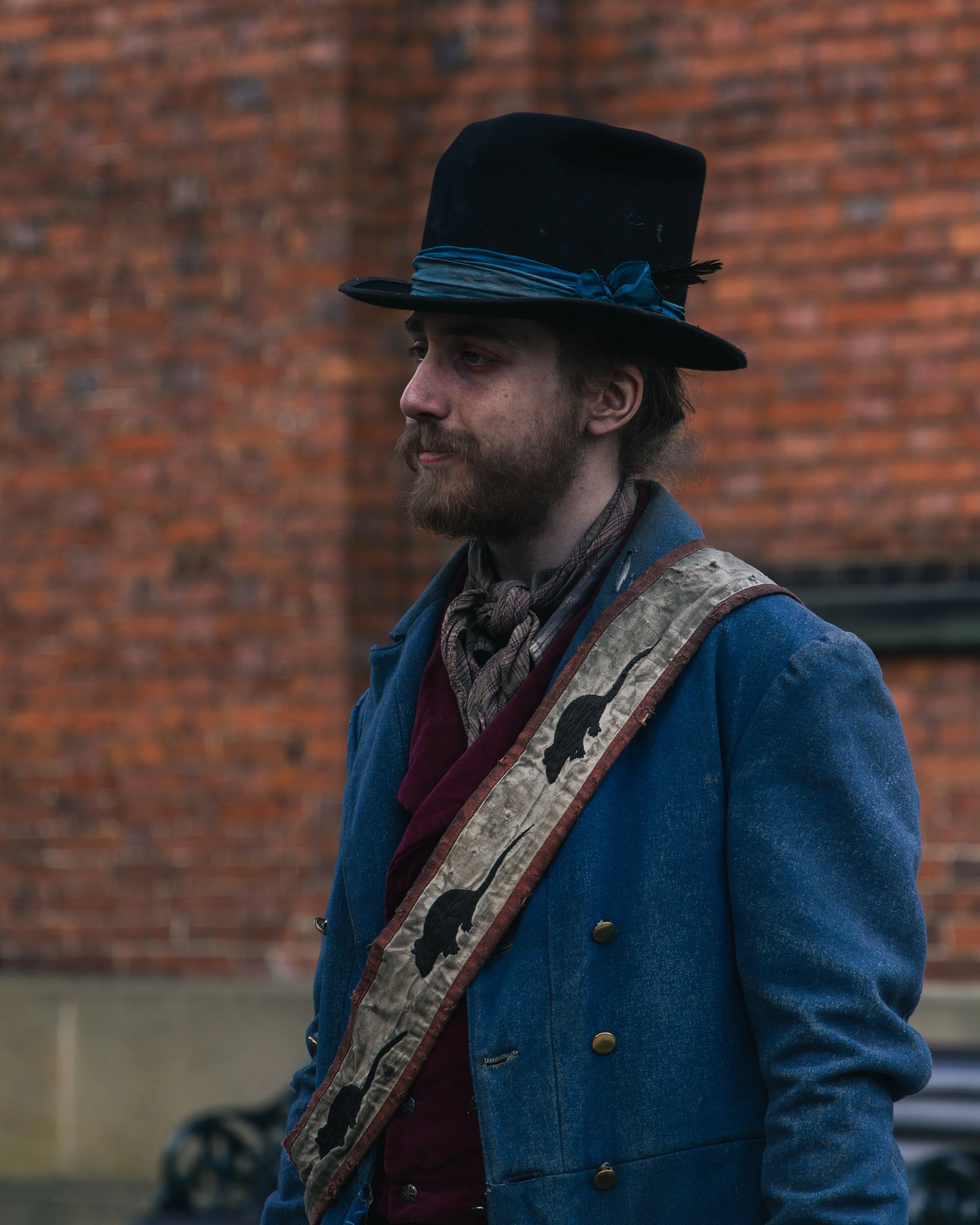 A man with a beard and long hair, wearing a black top hat with a blue scarf and feather, denim blue coat with brass buttons, walks beside a brick wall.