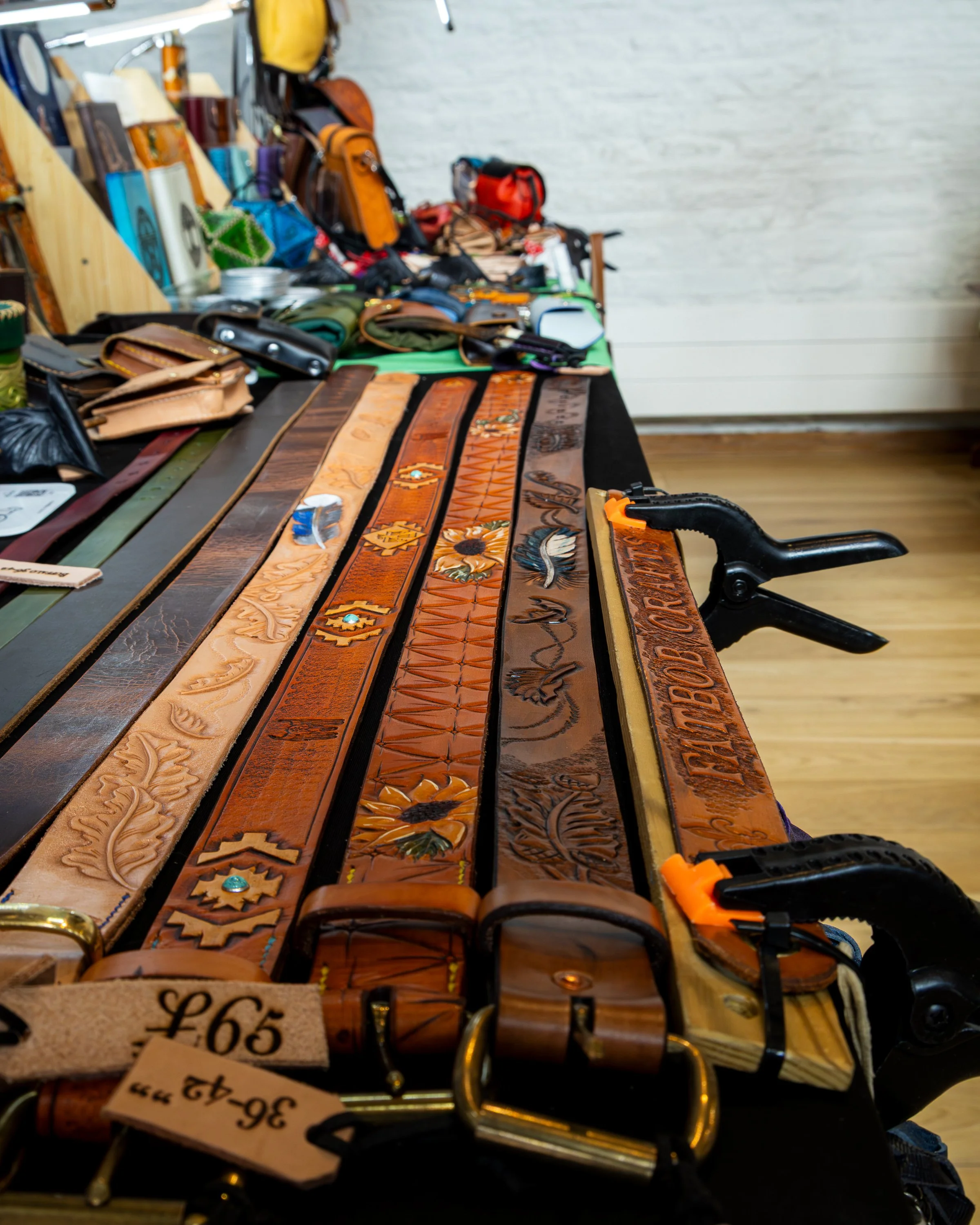 A table displaying various leather belts with intricate embroidery and designs, with some priced at 65 pounds. In the background, there are bags and other leather items on display.