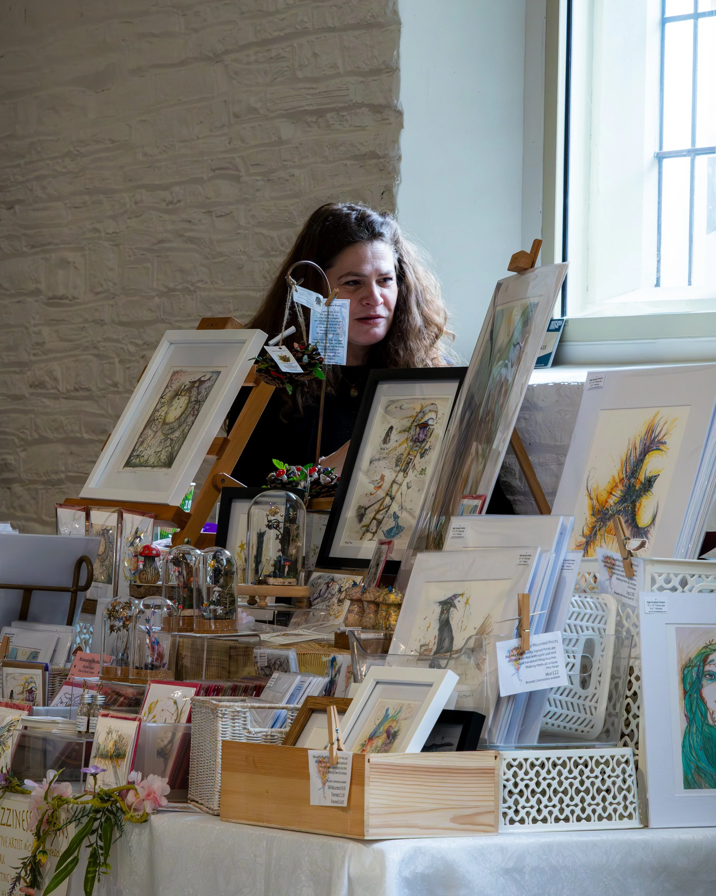 A woman browsing art prints at a craft fair or gallery, with artworks displayed on easels and a window in the background.