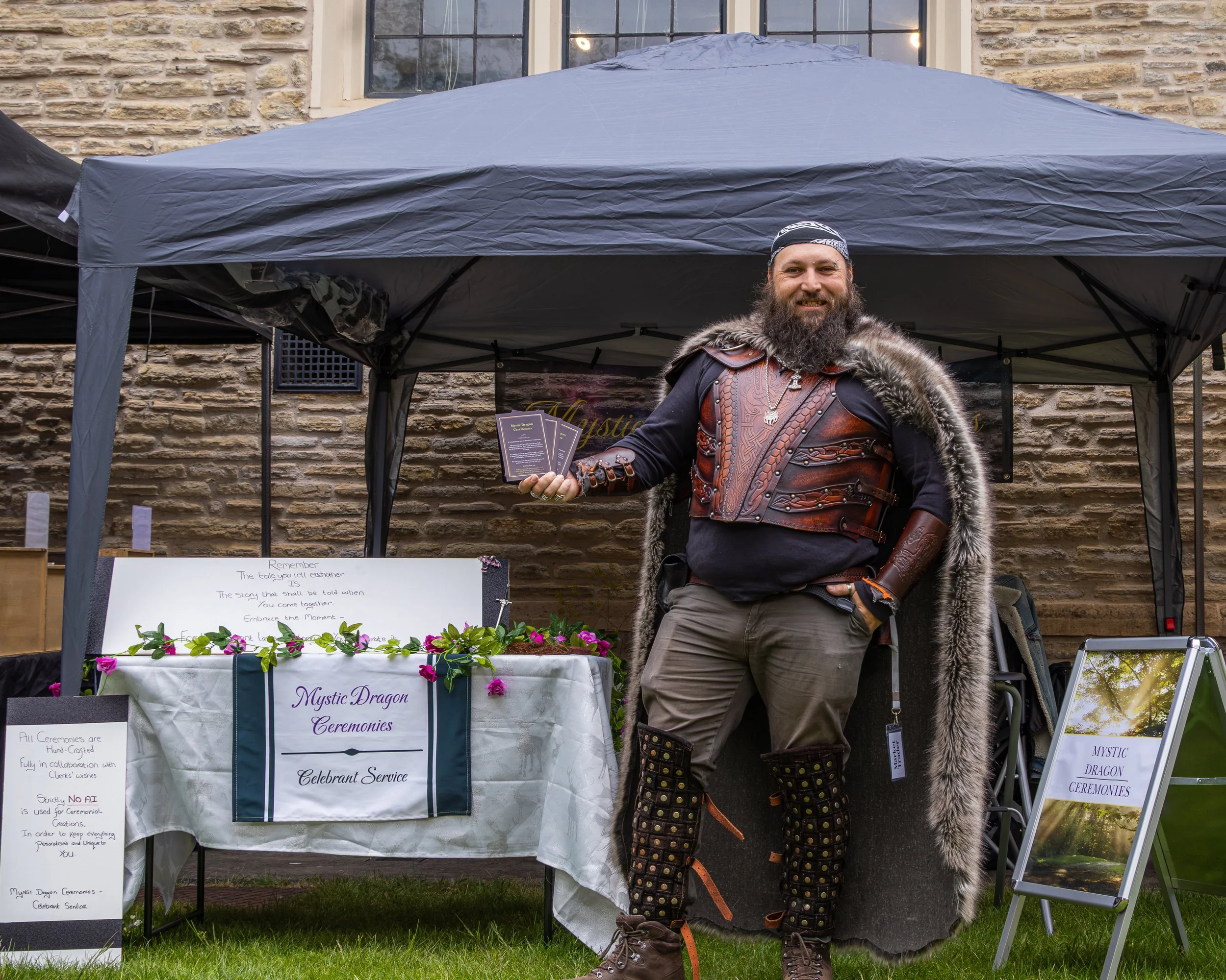 A man dressed in Viking-like costume with a fur cape, leather armor, patterned pants, and a bandana, standing outdoors in front of a booth labeled "Mystic Dragon Ceremonies" at a fair or event.