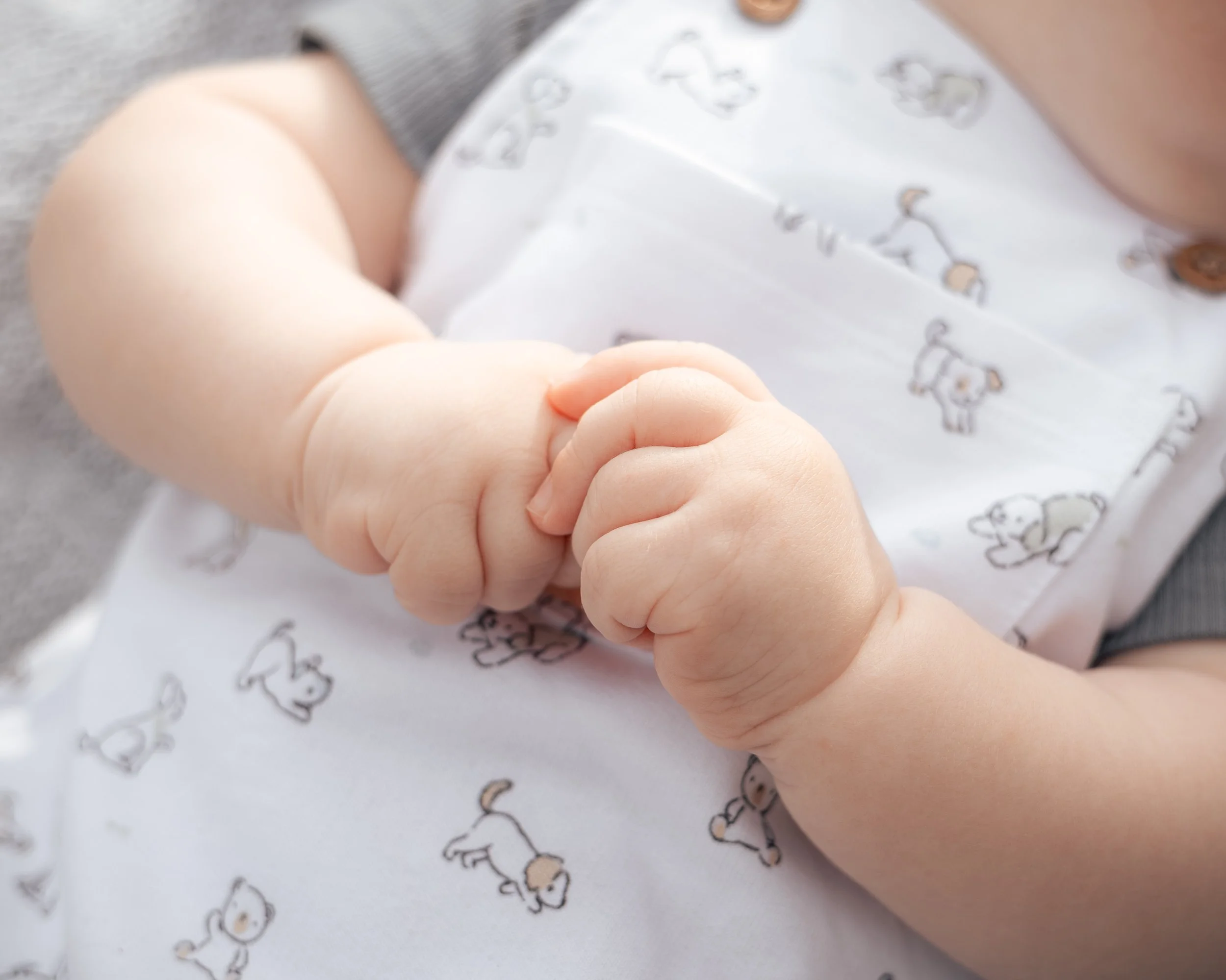 Close-up of a baby finger. The baby is wearing a white shirt with small animal prints. The background shows a soft surface with similar animal print designs.