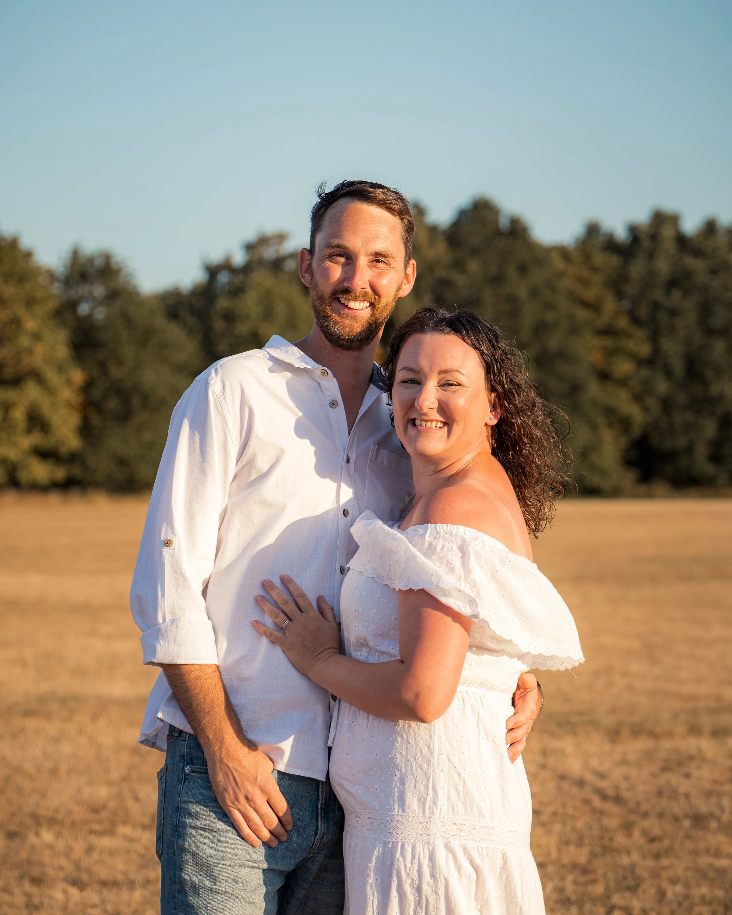 A smiling couple standing outdoors in a field during sunset, with the man wearing a white shirt and the woman in a white off-the-shoulder dress, embracing each other.