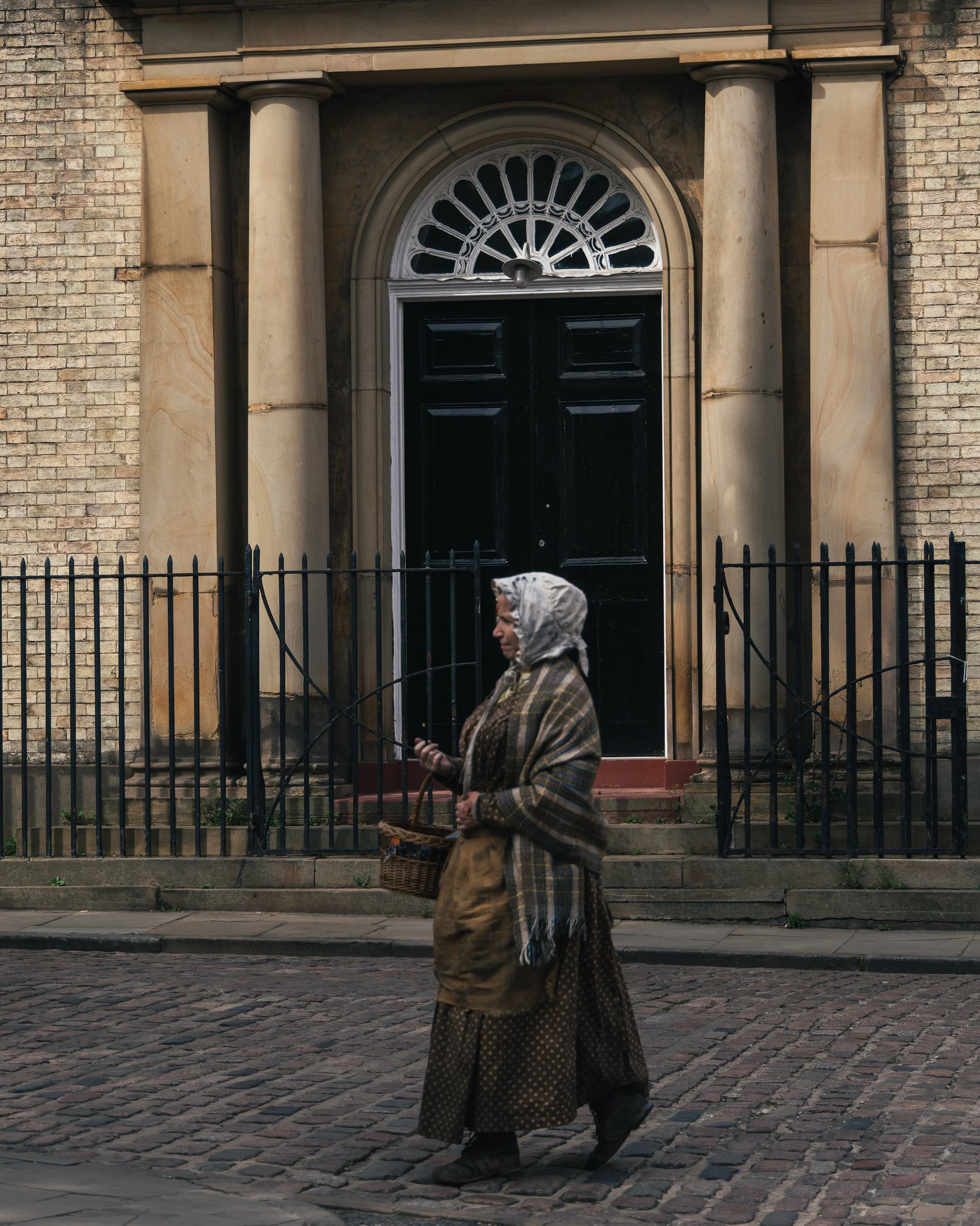 An elderly woman wearing traditional clothing, including a plaid shawl, a long skirt, and a headscarf, is walking past a black metal fence in front of a brick building with large columns and an arched window. She is holding a basket and looking down 