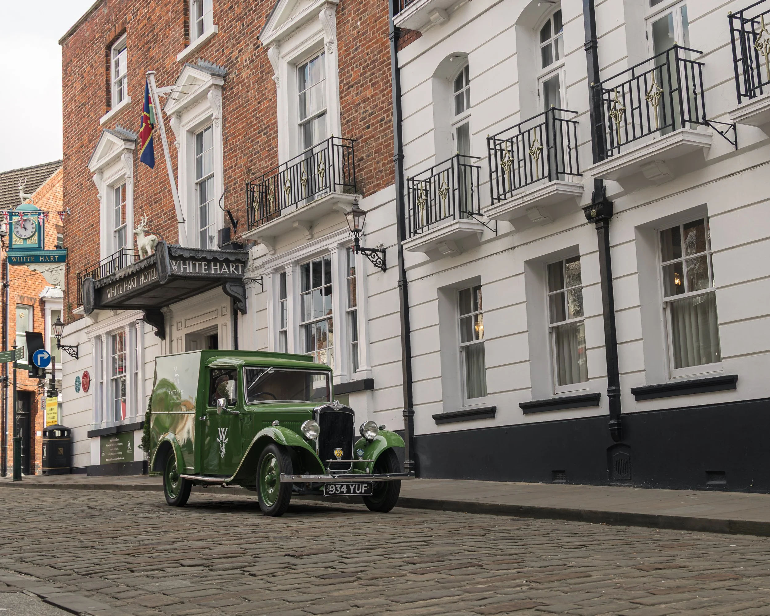 Vintage green car parked in front of a building with white and brick facade, black iron balconies, and a street sign for White Hart Hotel.