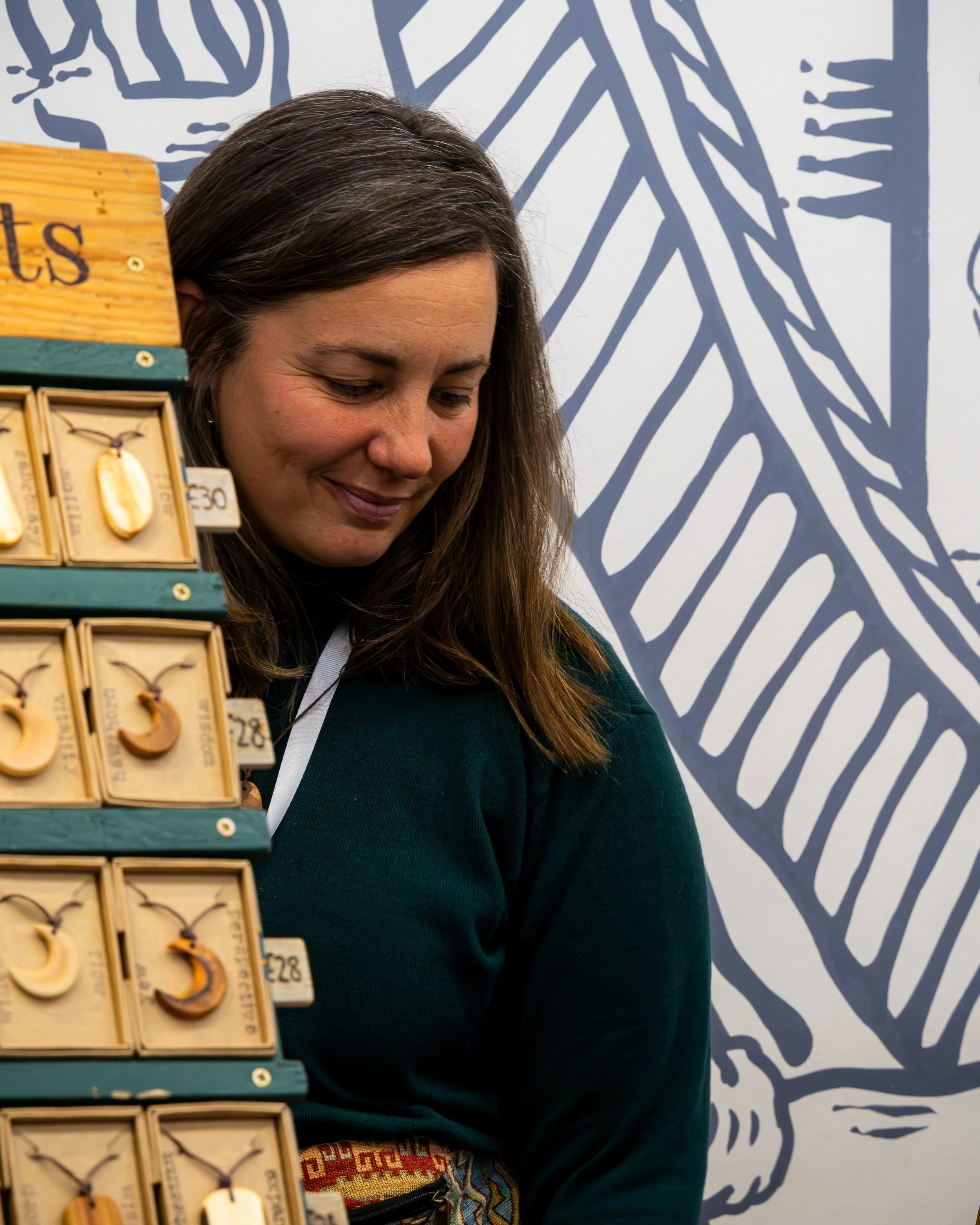 A woman with long brown hair smiling while looking down, standing next to a display of wooden jewelry tags with earrings on them, in front of a wall with a large blue and white geometric pattern.