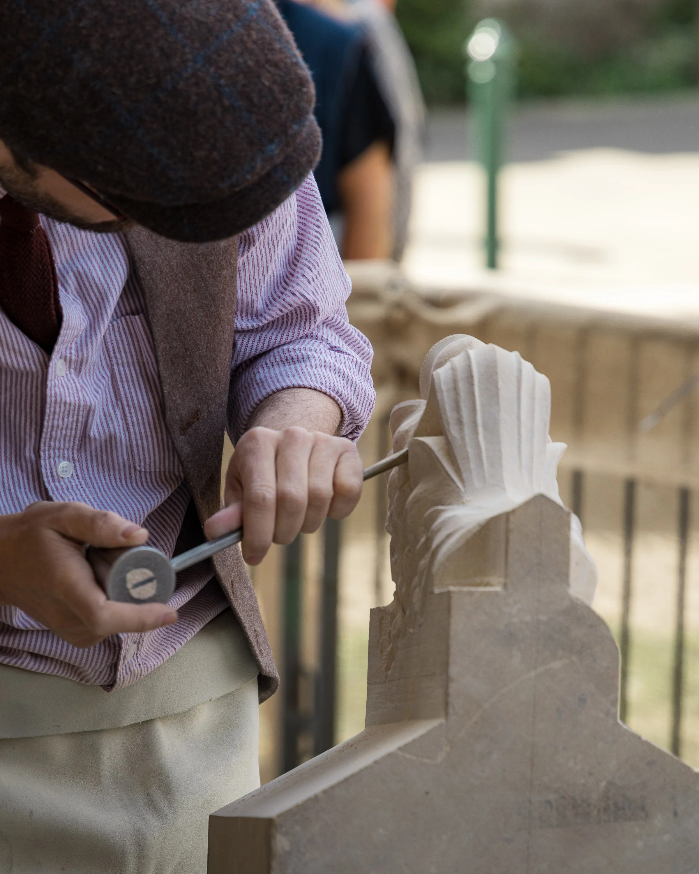 Close-up of a person carving a wooden sculpture with a chisel and hammer. The individual is wearing a brown wool hat, a red and white striped shirt, and a grey vest. The focus is on the hands and the detailed wooden figure they are working on.