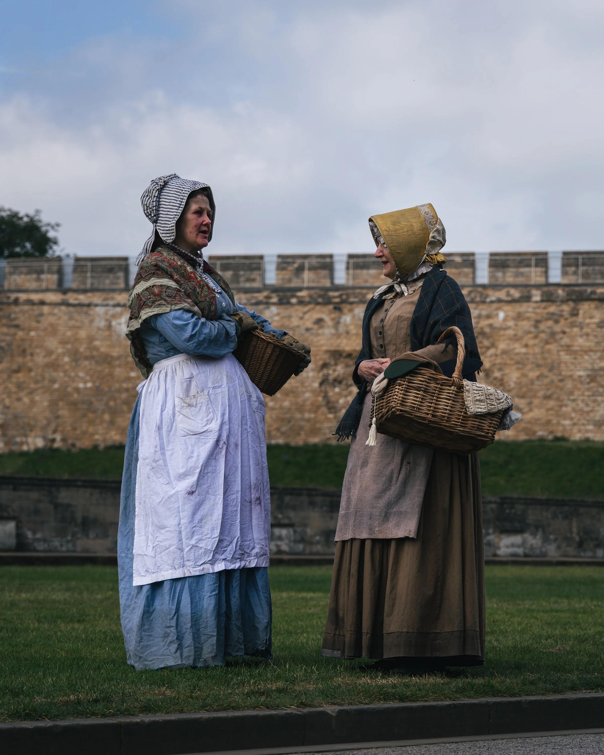 Two women dressed in historical clothing, wearing bonnets and shawls, standing outdoors in front of a stone wall, engaged in conversation, each holding a wicker basket.