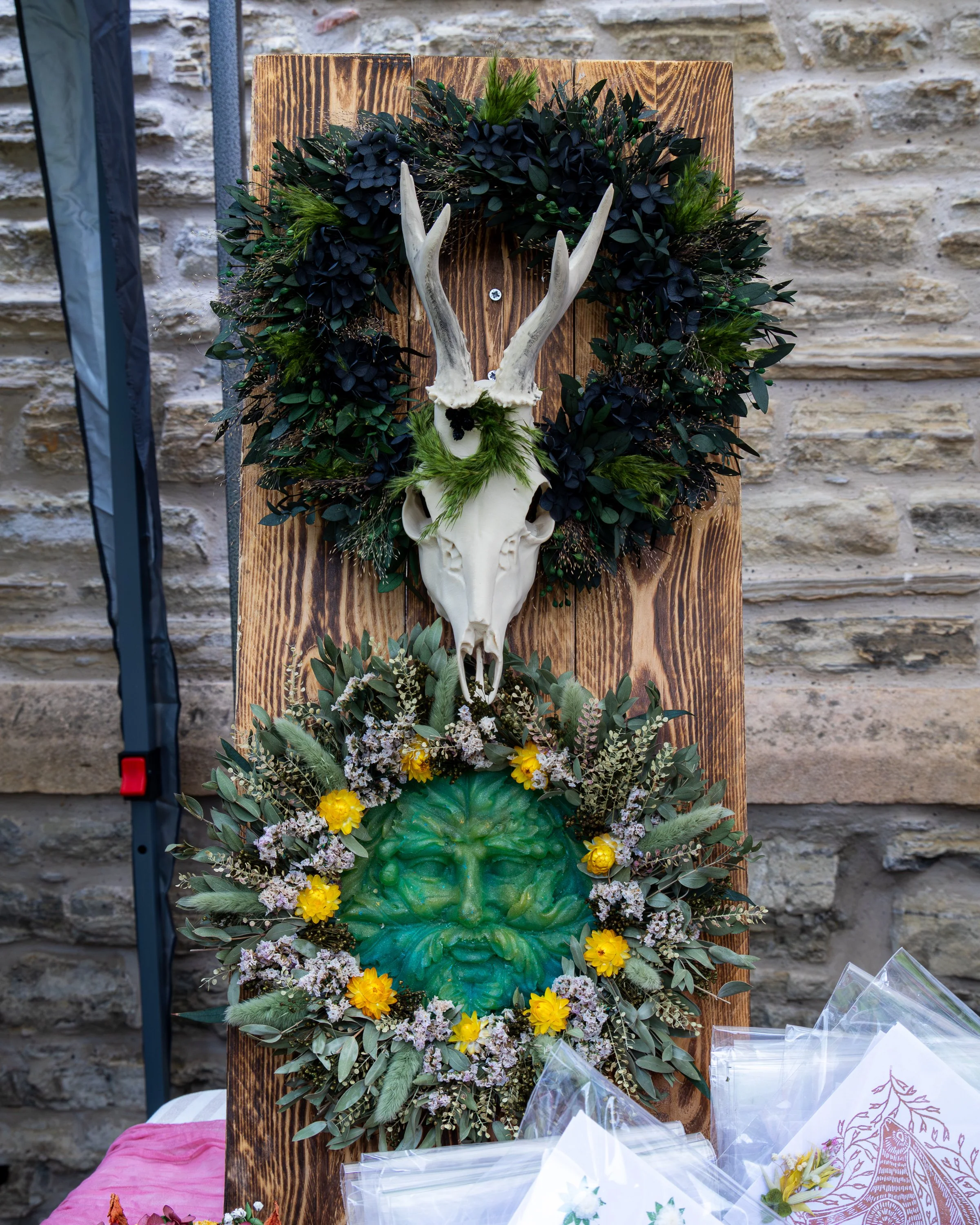 Decorative display featuring a skull with antlers, surrounded by lush greenery and yellow flowers, mounted on a wooden panel against a stone wall background.
