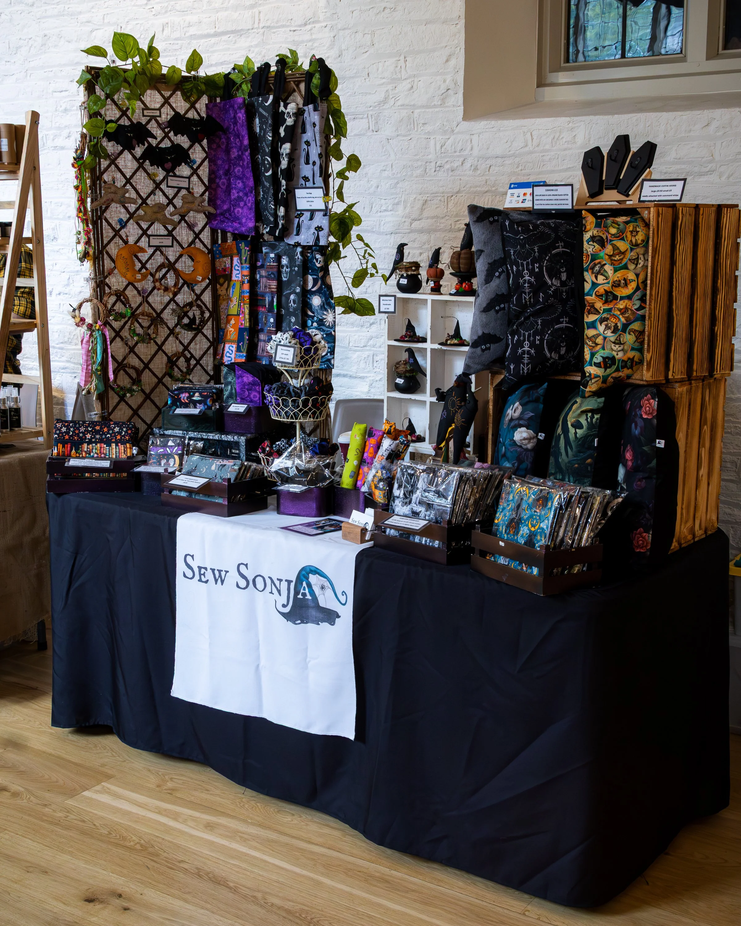 A display table with black and white fabric items, including bags, scarves, and pouches, set up at a craft fair or market. The table is covered with a black cloth and has a sign that reads 'Sew Sonja'. Behind the table, there is a white brick wall wi