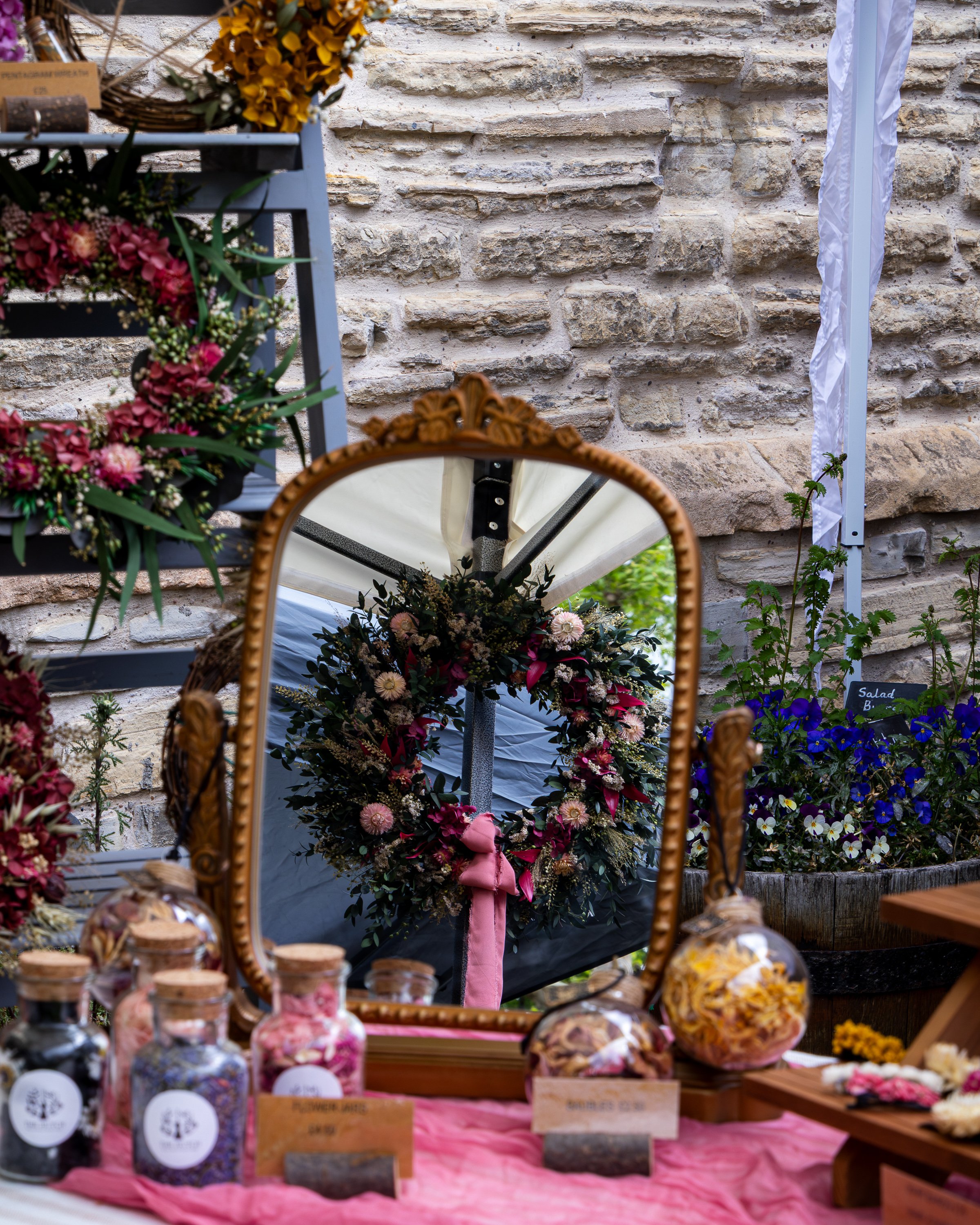 A display of colorful flower arrangements and glass jars filled with various treats at an outdoor market stall against a stone wall background.