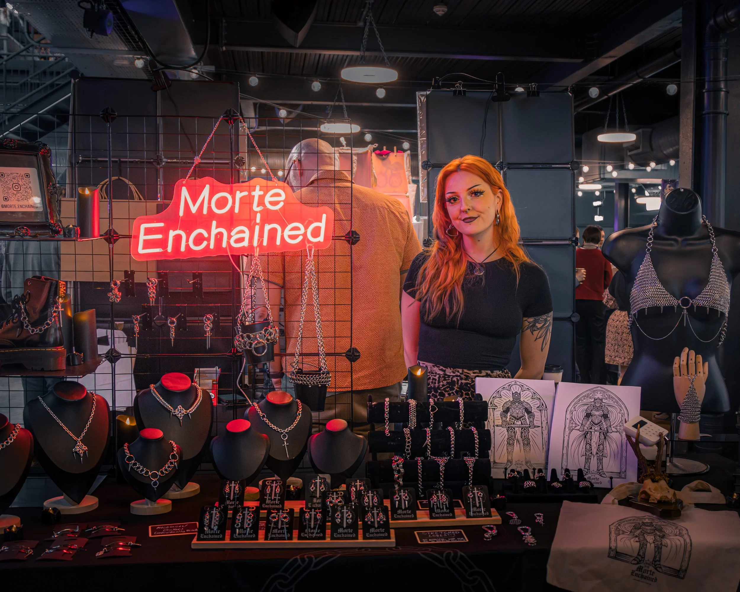 A woman with red hair, wearing earrings and dark lipstick, stands behind a jewelry display at a booth. The booth has a red neon sign that says 'Morte Enchained' and showcases various silver jewelry, including necklaces, bracelets, and rings, on black