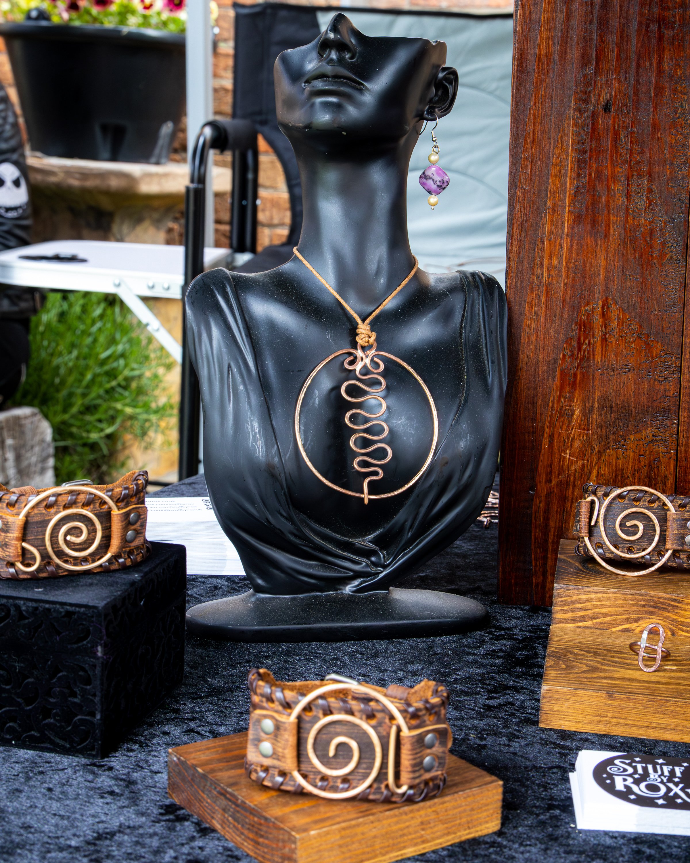 A black mannequin bust displaying jewelry, including a gold necklace with a knot and intricate wire design, an earring with a purple gemstone and pearl, and a wooden bracelet with metal spiral accents. The display is set outdoors with other jewelry i