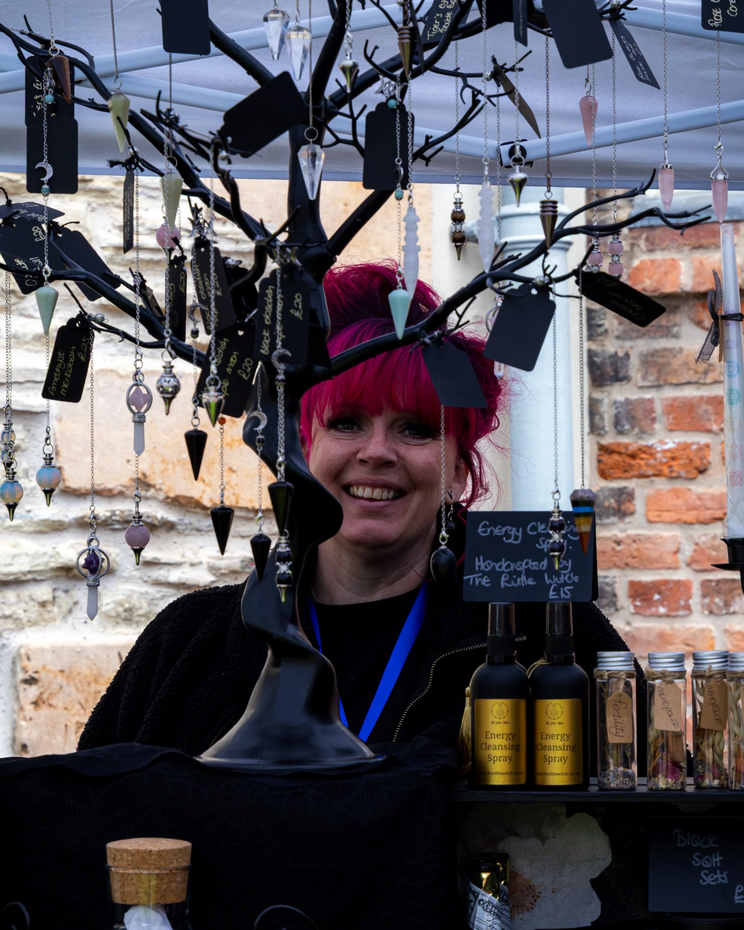A person with bright pink hair smiling behind a display of necklaces and bottles labeled 'Energy Cleansing Spray'. The display is set against a brick wall.