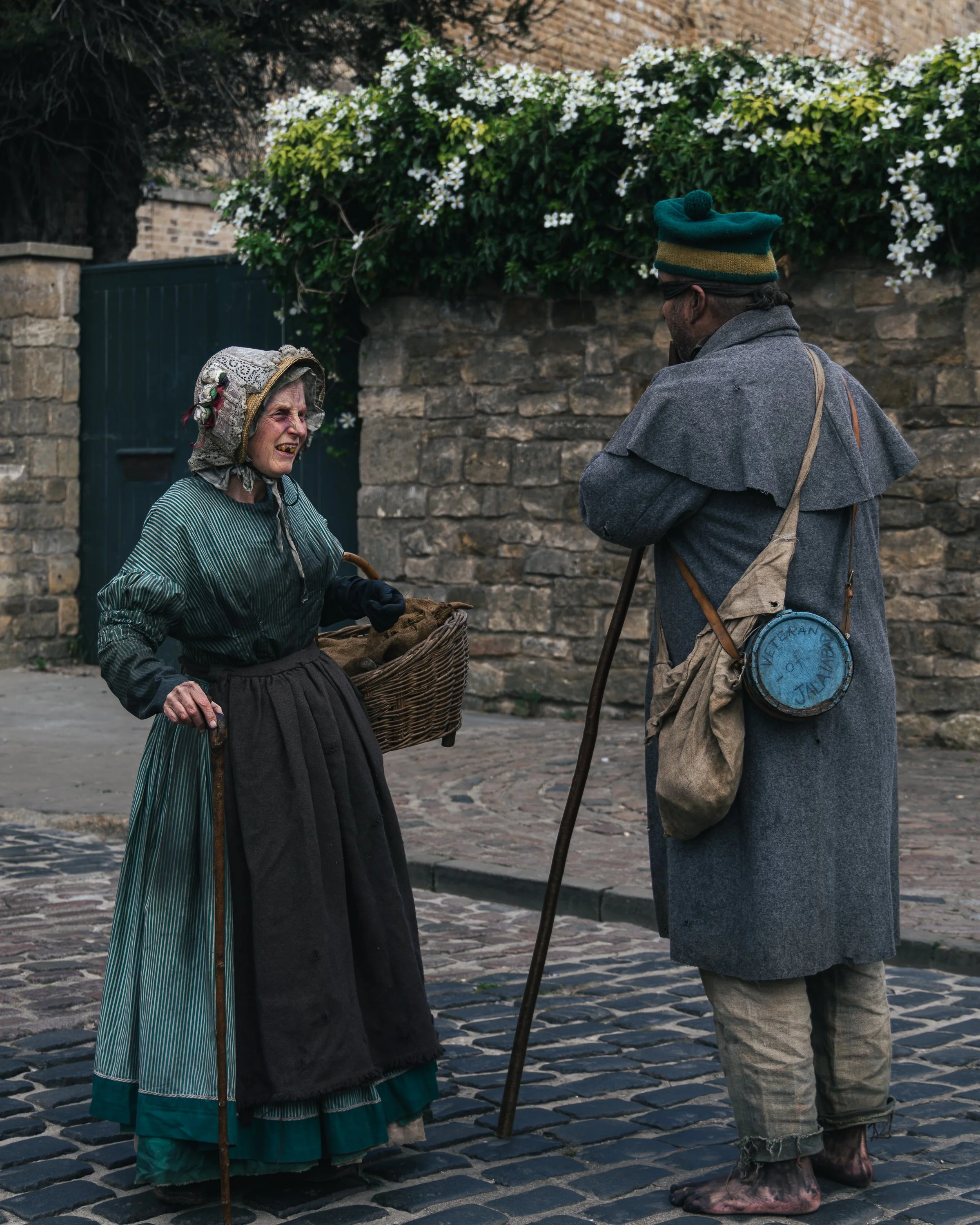 An elderly woman and an elderly man are talking to each other outdoors on a cobblestone street. The woman is dressed in traditional clothing with a headscarf, and she holds a cane and a basket. The man is wearing a coat, a hat with a pom-pom, and has