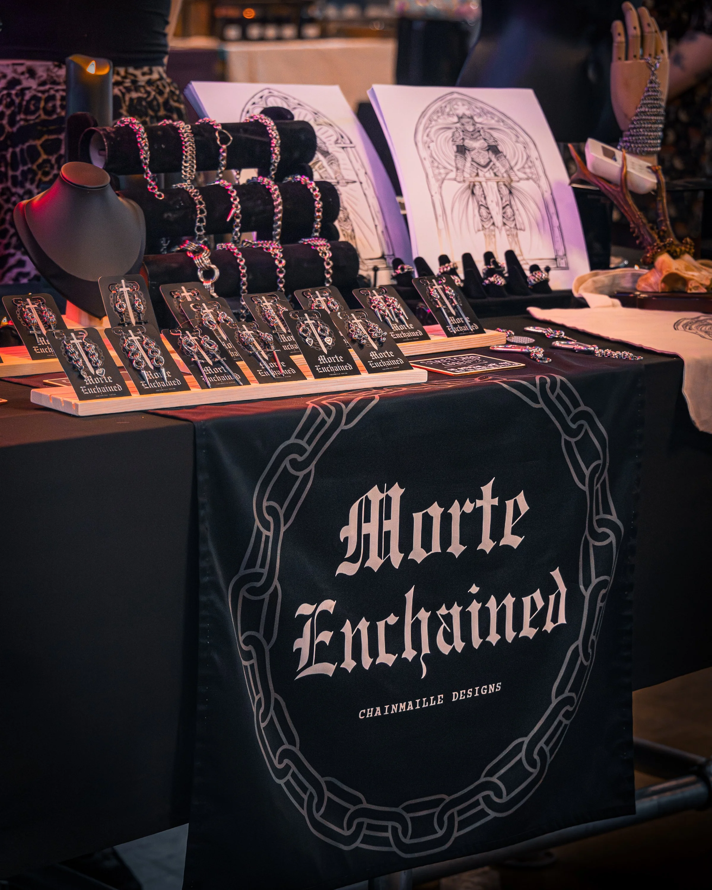 Jewelry display at a market stall, featuring silver chains, earrings, and artwork with a medieval knight design, with a black tablecloth that has a chain-link pattern and a gothic-style logo.