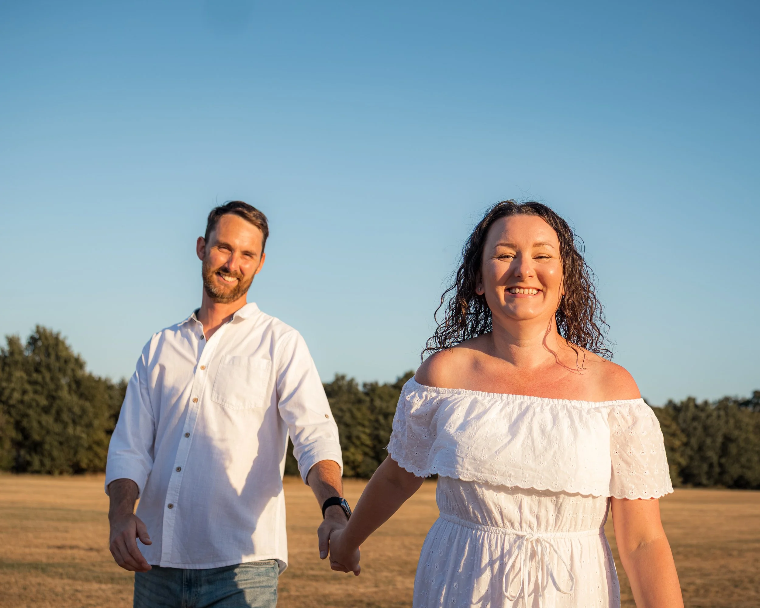 A smiling woman in a white off-shoulder dress holding hands with a man in a white shirt and jeans, walking outdoors during daylight with a clear blue sky and trees in the background.