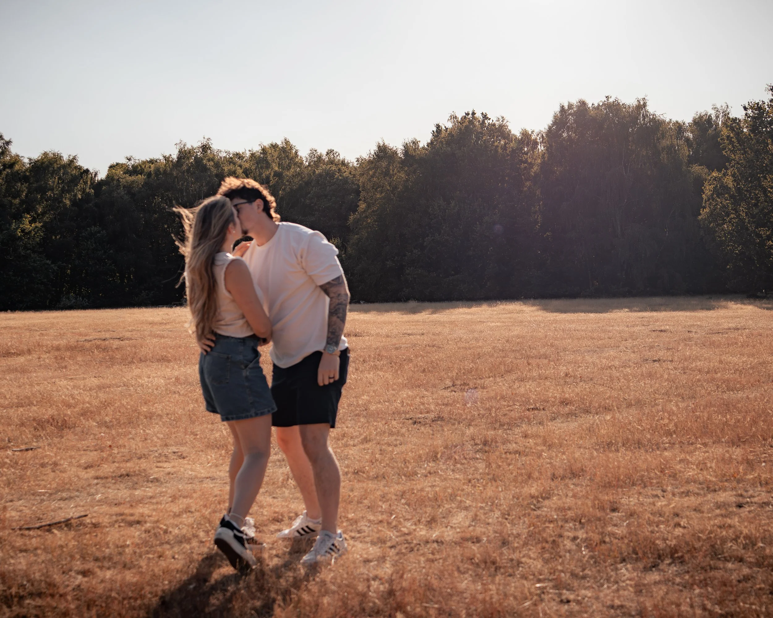 A couple kissing in a field with trees in the background during sunset.
