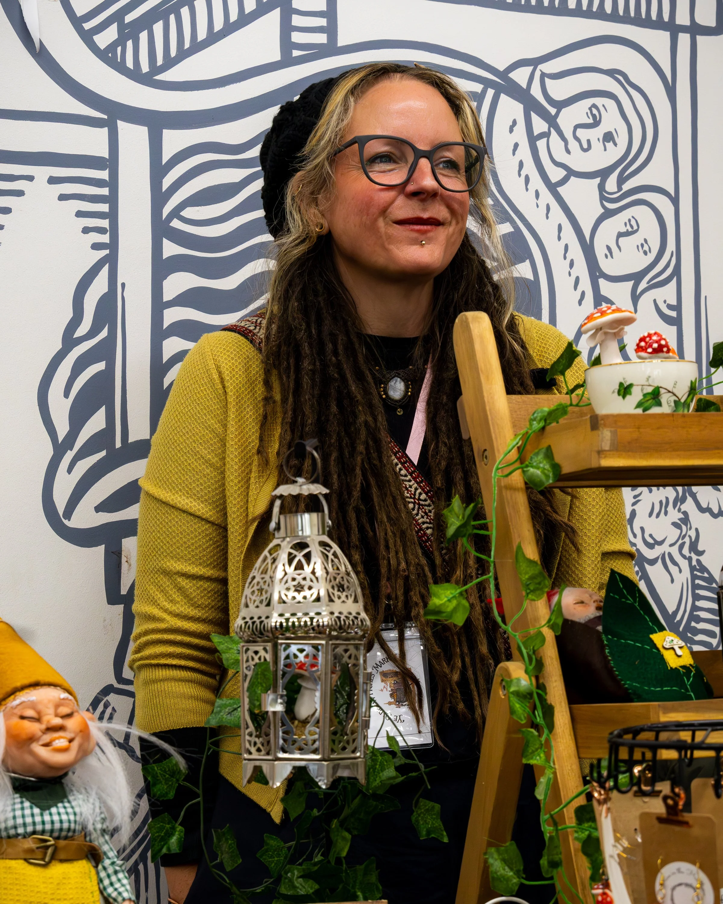 A woman with light skin, glasses, and long dreadlocks smiling at a craft fair or display, with decorative mushrooms and plants on display in front of her, and a mural with abstract lines in the background.