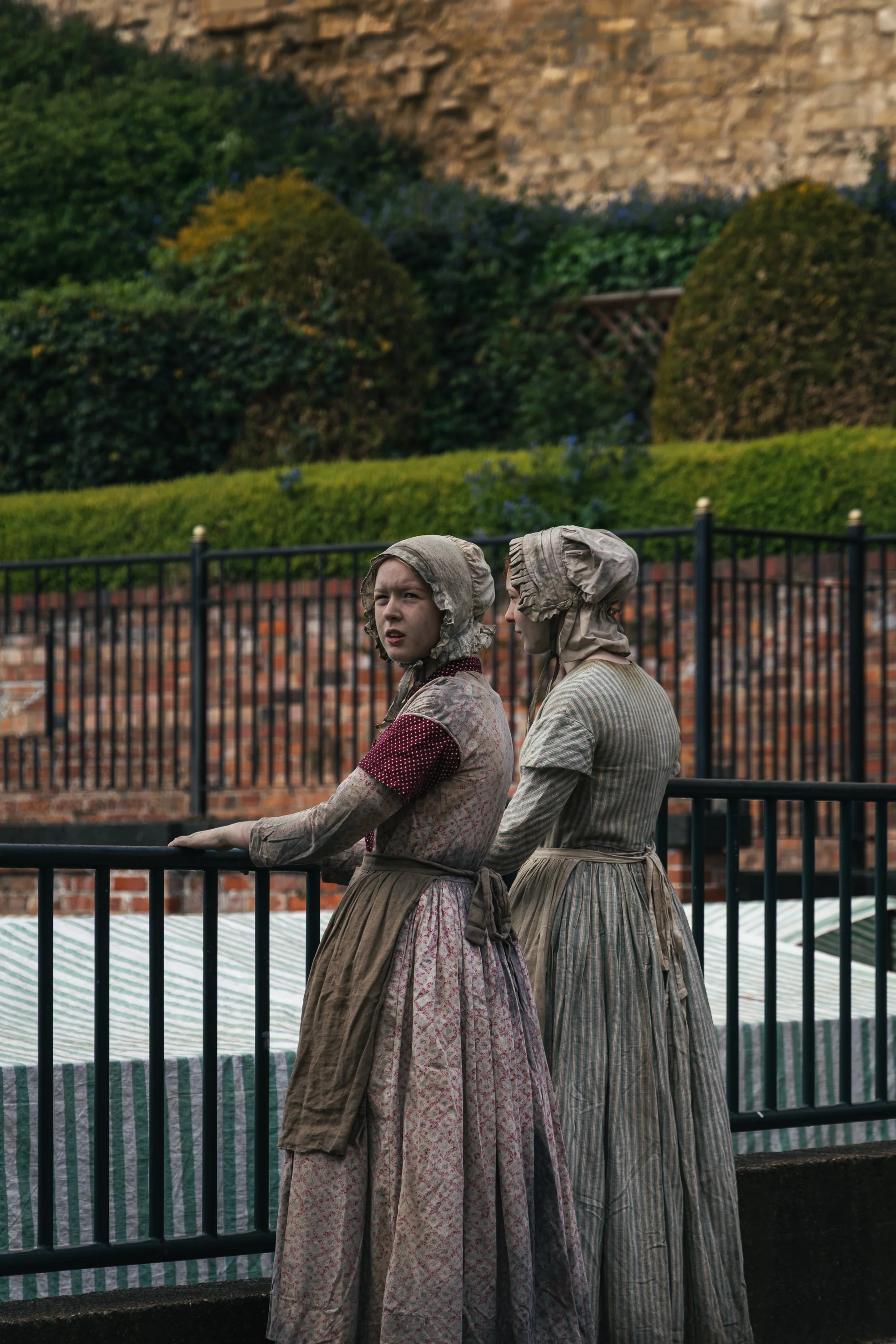 Two women dressed in 18th-century period clothing, wearing bonnets, standing behind a black metal fence with a brick wall and greenery in the background.