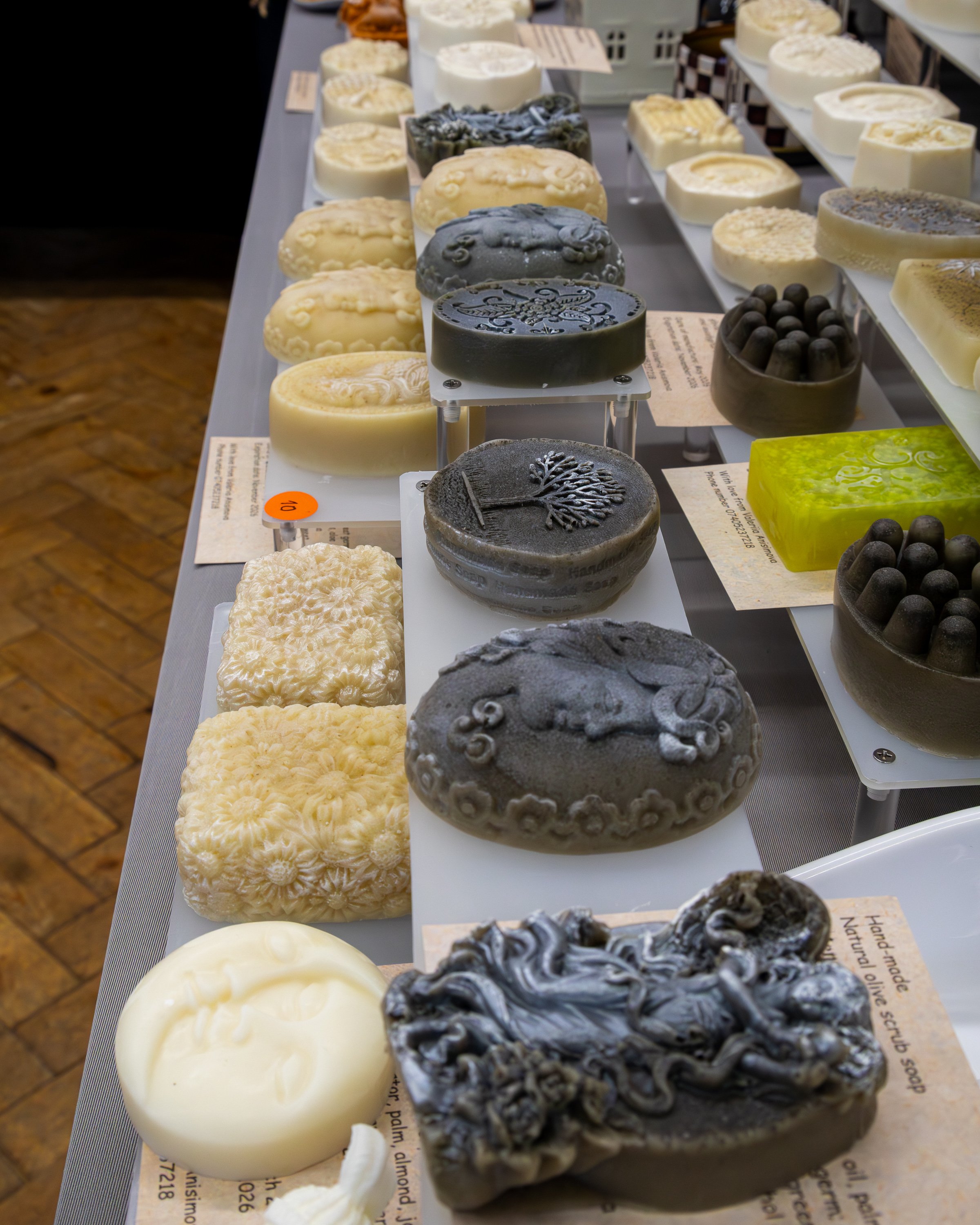 Display of decorative soaps at a market or artisan shop, arranged on white shelves, with various colors and textures, including black, cream, and marbled designs.