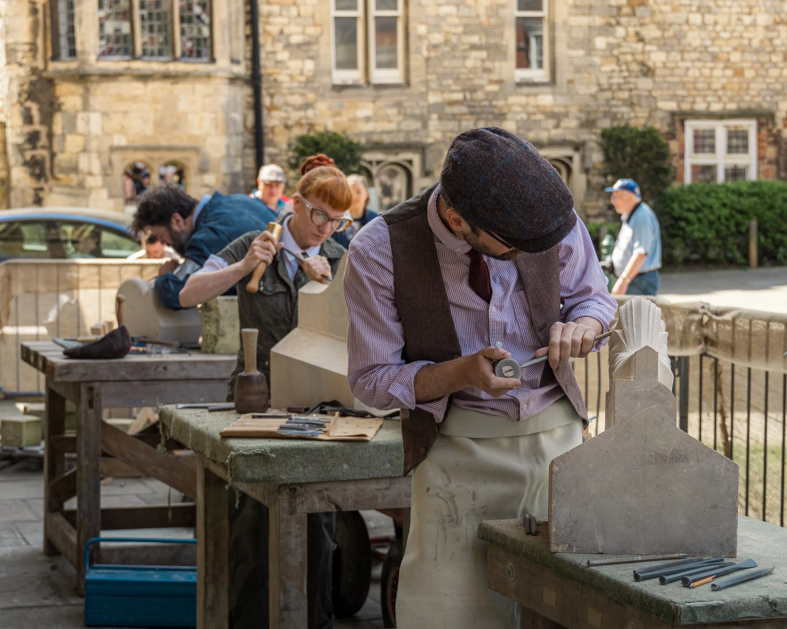 A man with a flat cap and glasses carving a stone sculpture outdoors, with other people working on similar projects behind him, and a brick building in the background.