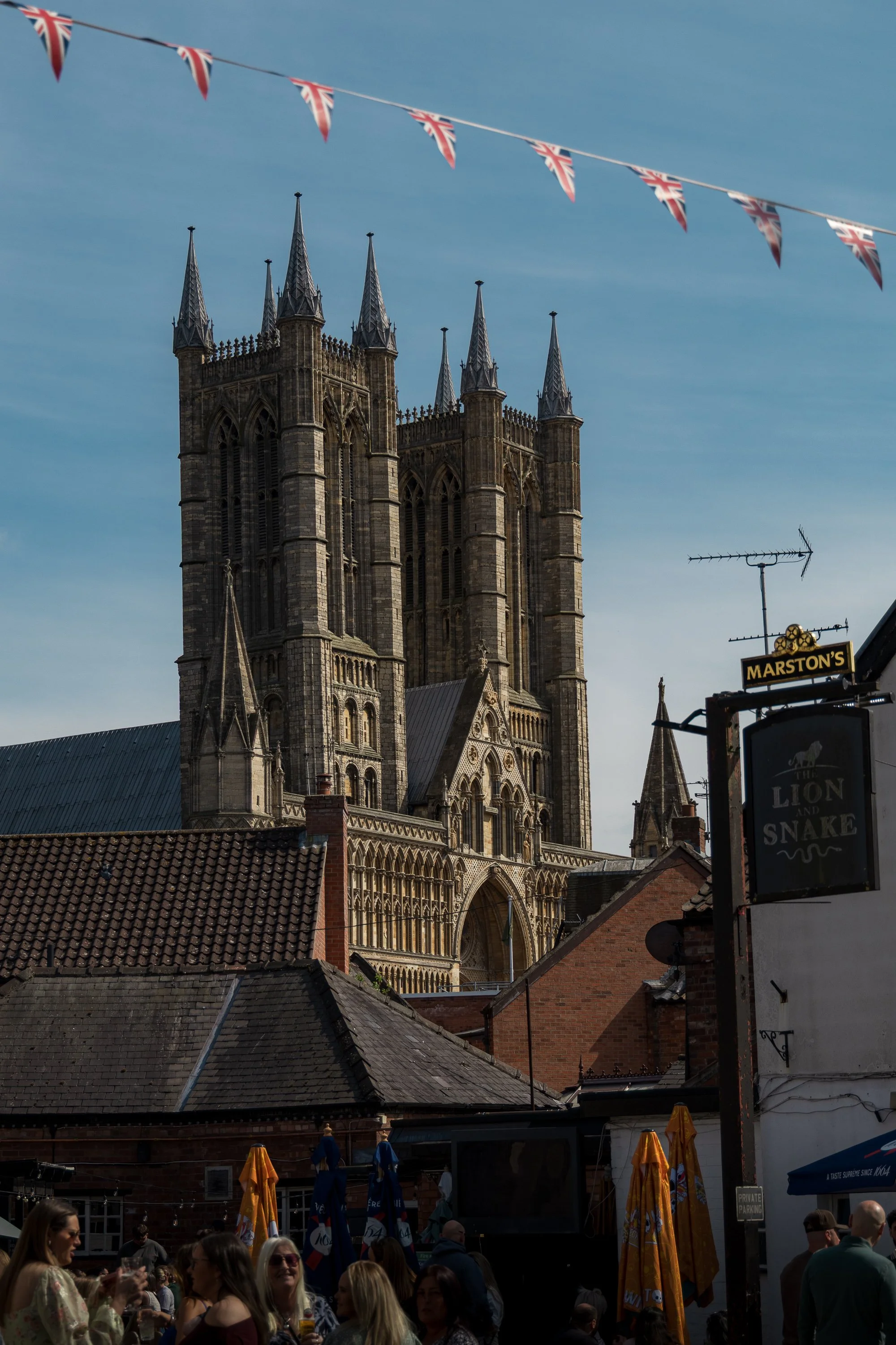 Crowd of people gathered outdoors near a market stall with yellow umbrellas, with a historic stone church with tall spires in the background, under a clear blue sky with Union Jack flags hanging above.