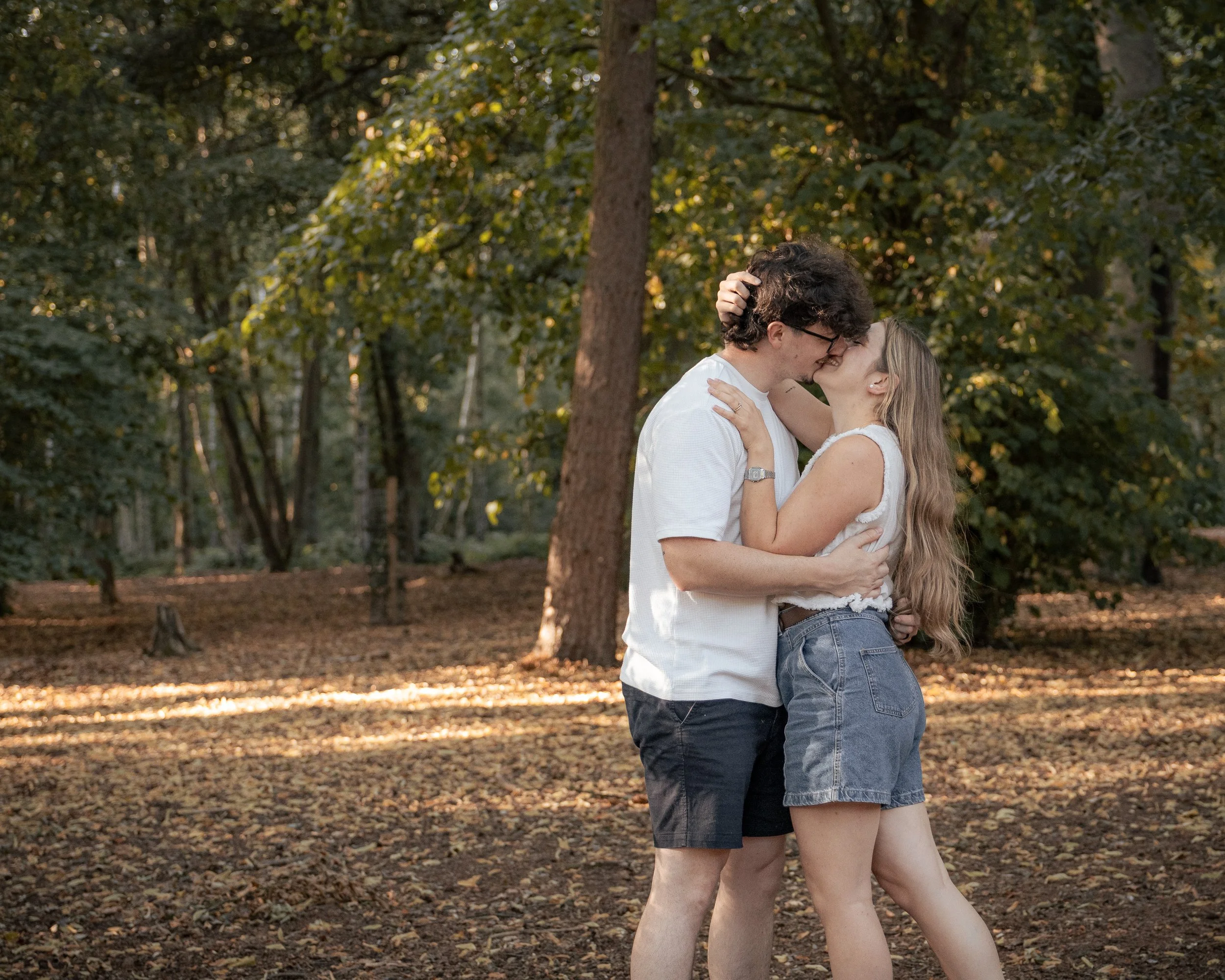A couple sharing a kiss in a park surrounded by trees with autumn leaves.